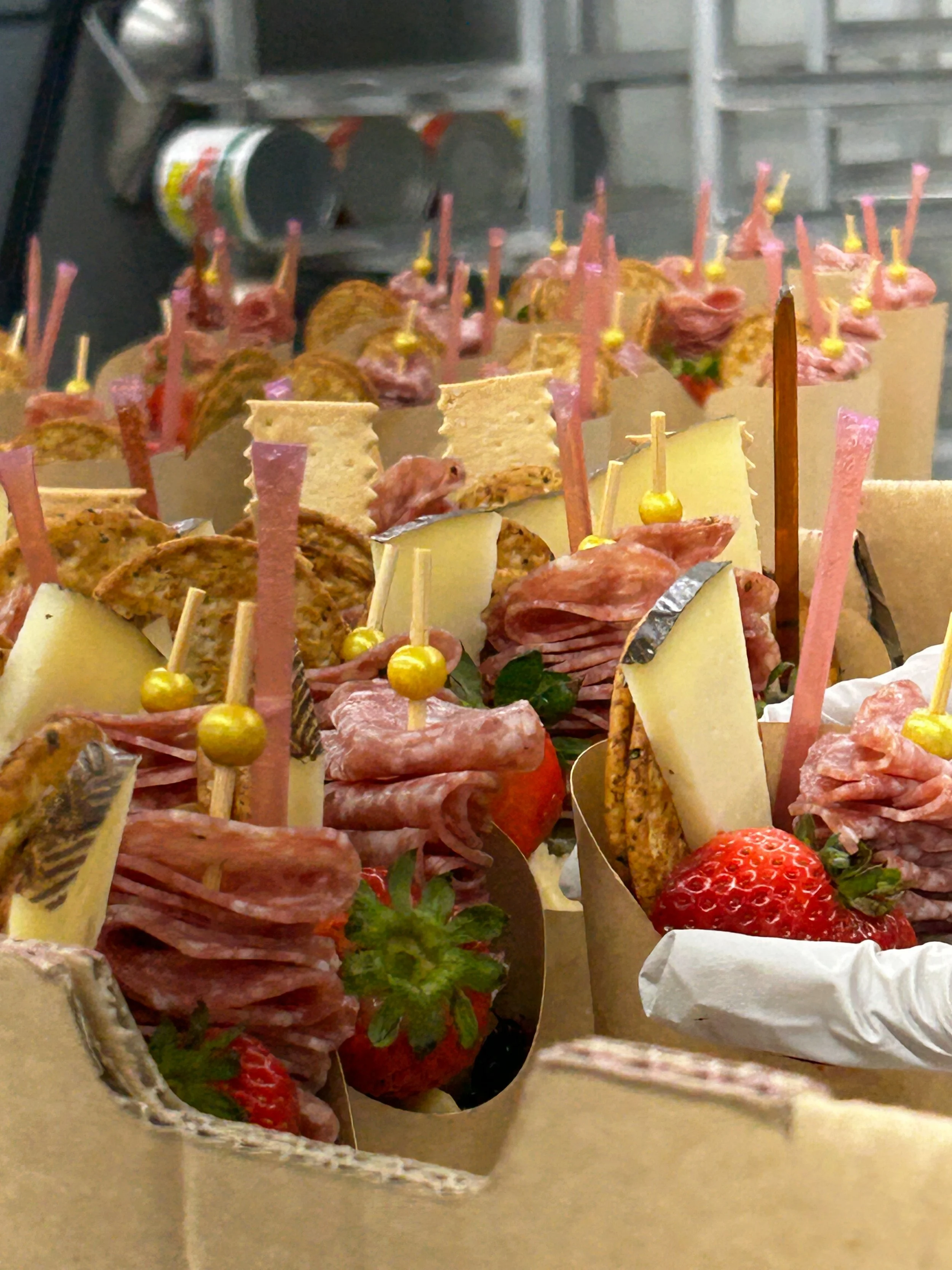 Skewers with slices of cured meat, cheese, strawberries, and garnishes in a cardboard container.