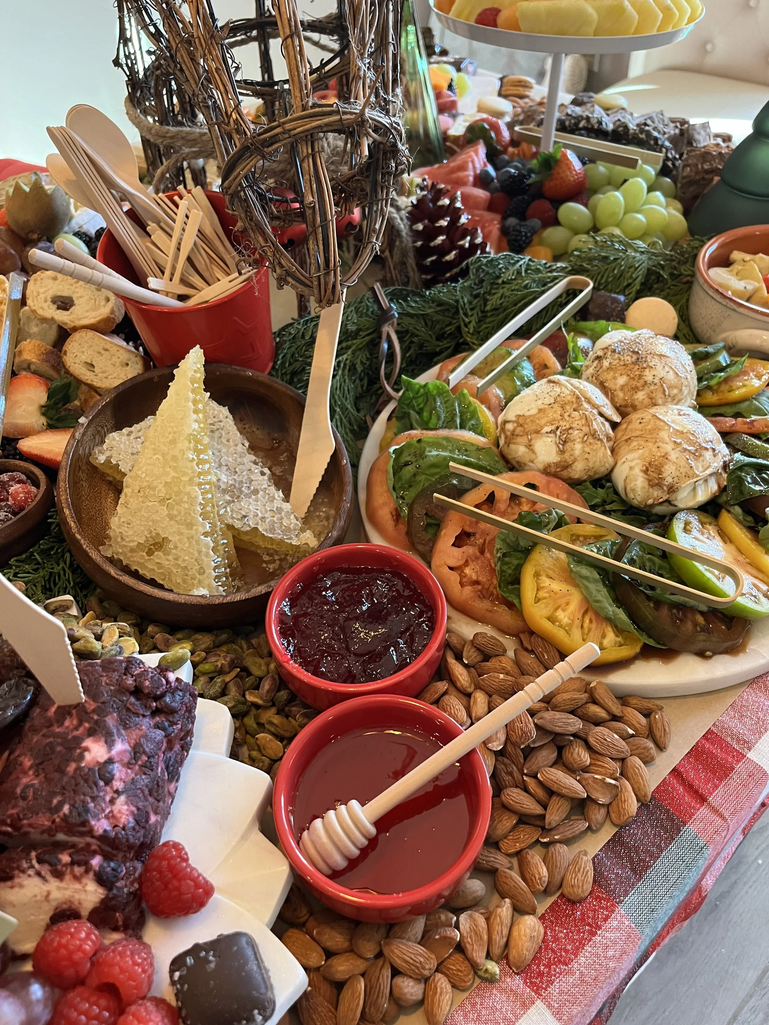 A festive food display with honeycomb, red fruit jam, almonds, roasted vegetables, grapes, strawberries, watermelon, chocolate dessert, and various bowls of fruit and sweet treats on a table with a red and white checkered tablecloth.