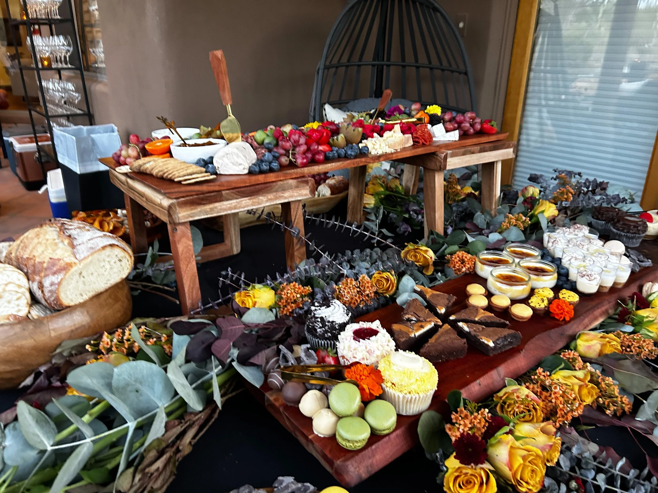 A spread of food on a table including bread, cookies, cakes, cheese, grapes, berries, and desserts, decorated with flowers and foliage.