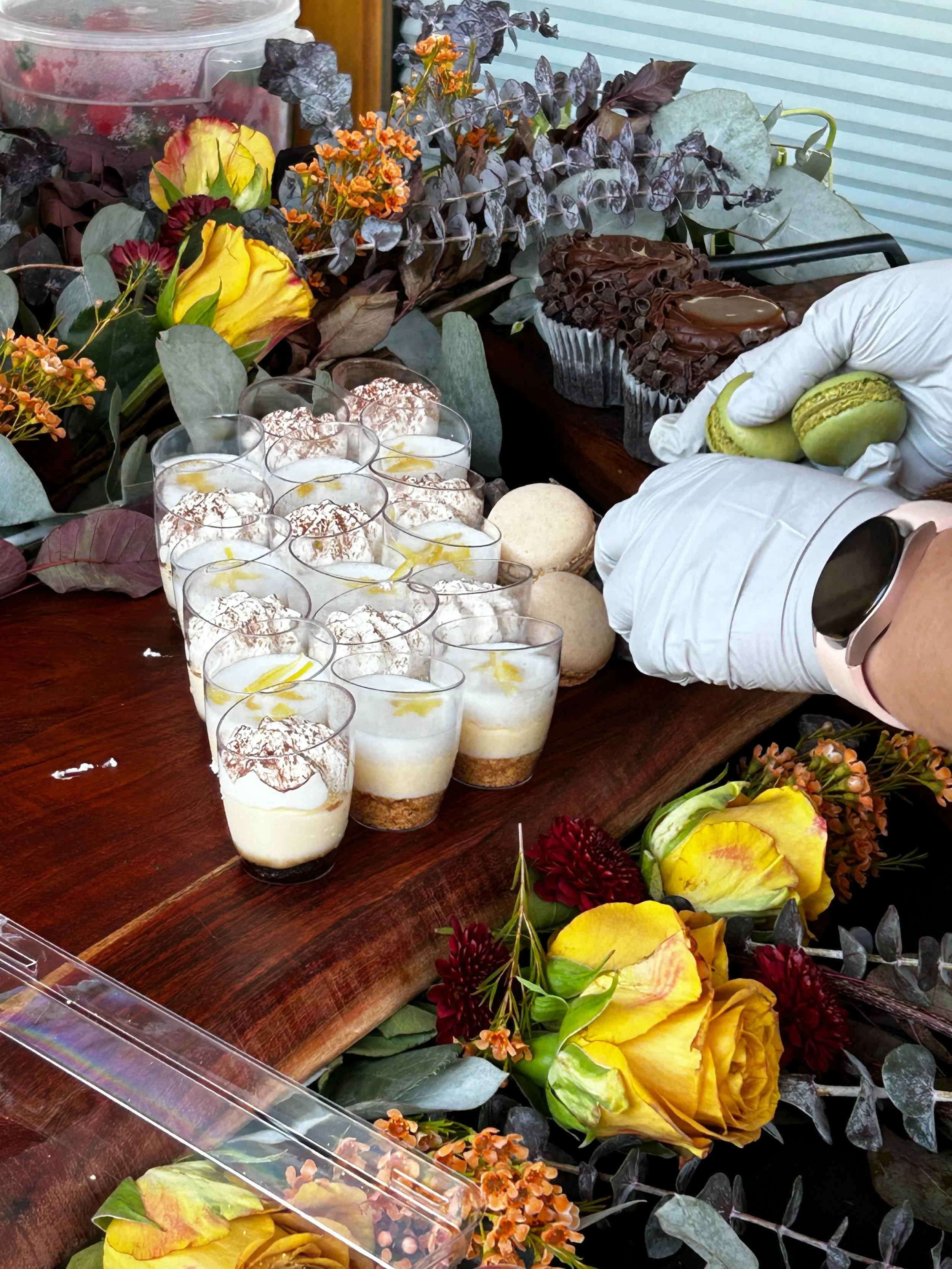 A person in white gloves decorating a dessert on a wooden table surrounded by flowers and desserts.