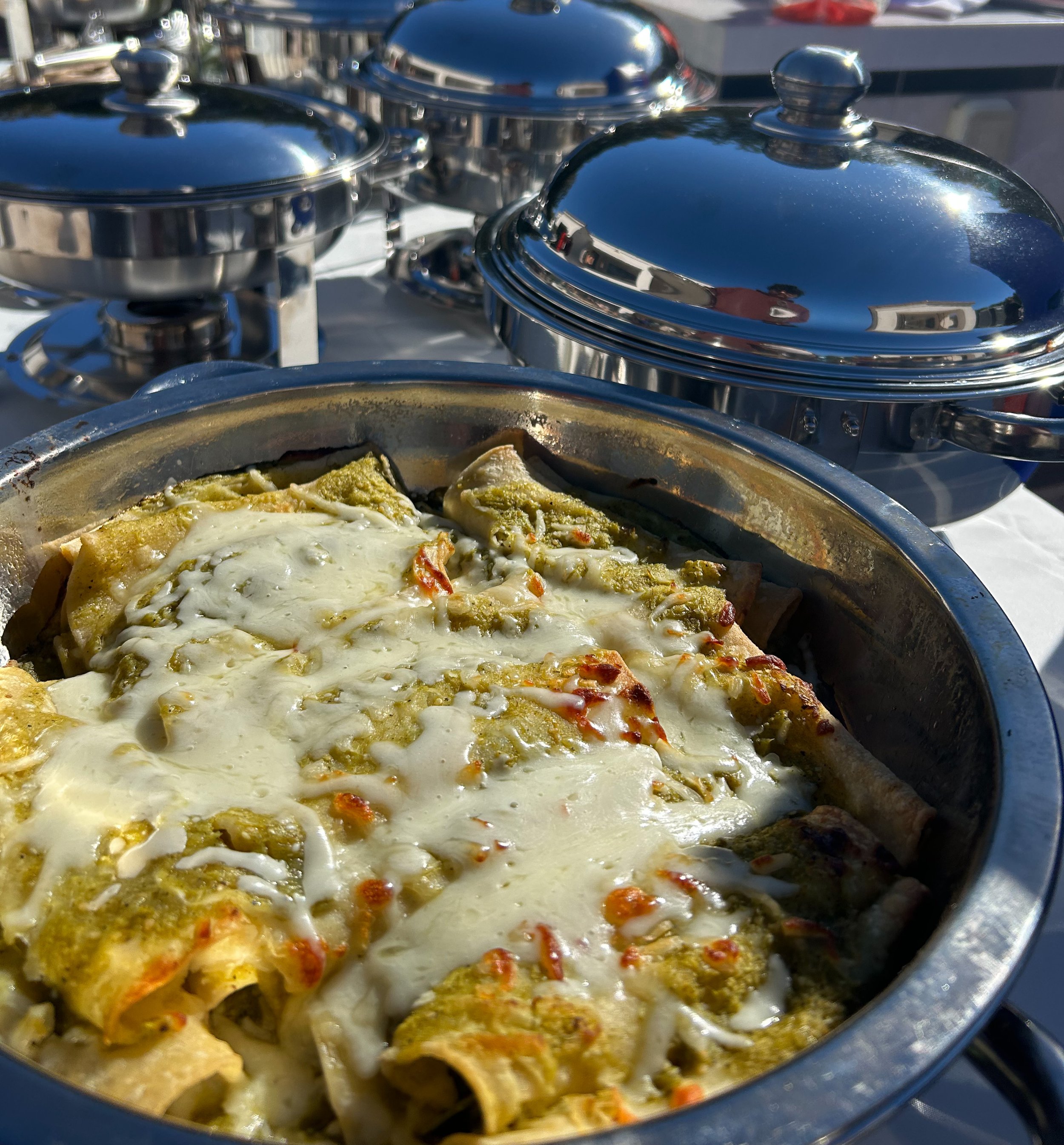 A metal chafing dish of baked pasta with melted cheese, set outdoors with several shiny metal chafing dishes in the background.