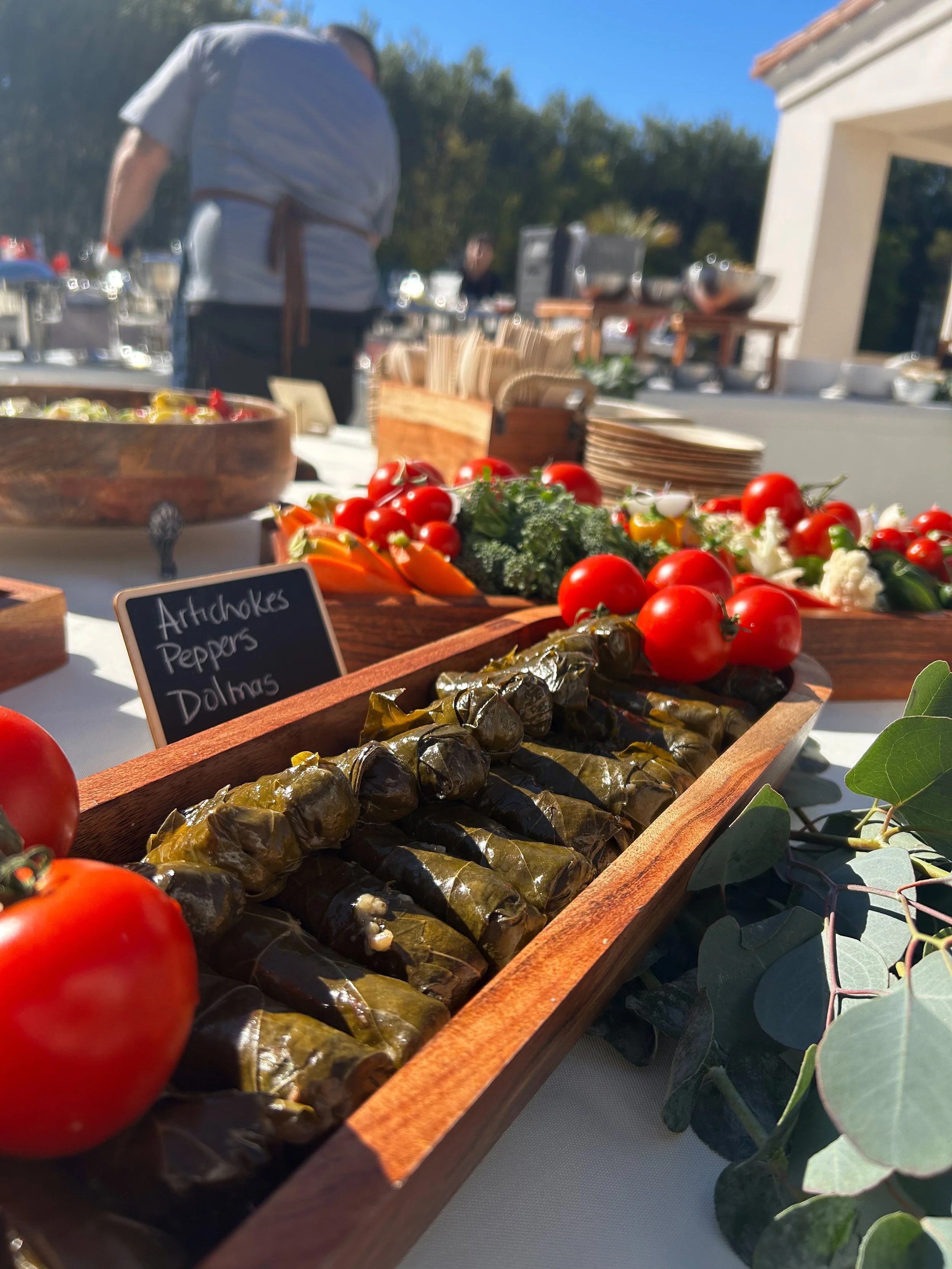 Wooden tray of stuffed grape leaves with cherry tomatoes, and other vegetables on a table at an outdoor buffet.
