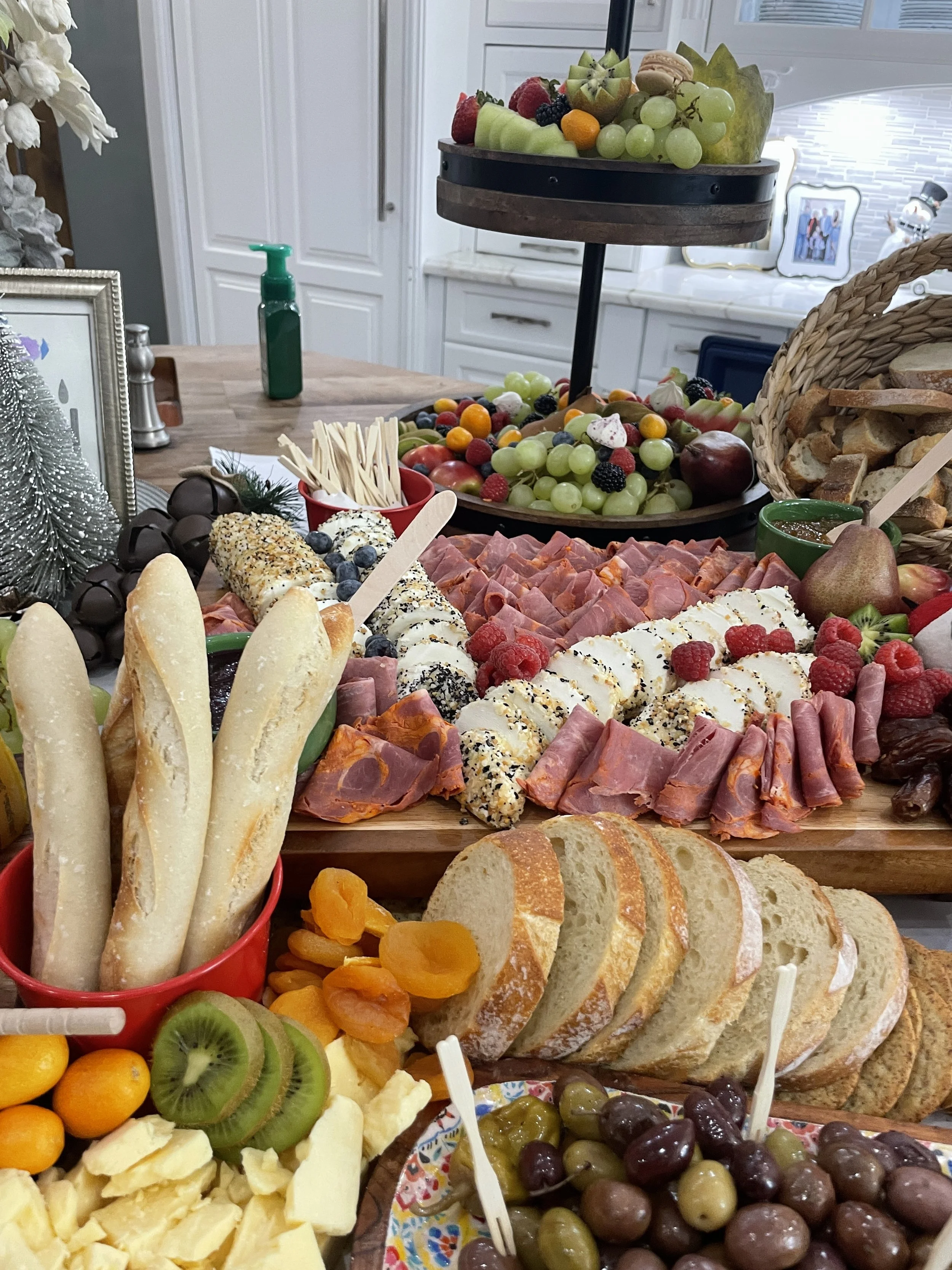 A buffet table with assorted breads, cheeses, sliced meats, fresh fruits, crackers, and olives, arranged for a gathering.