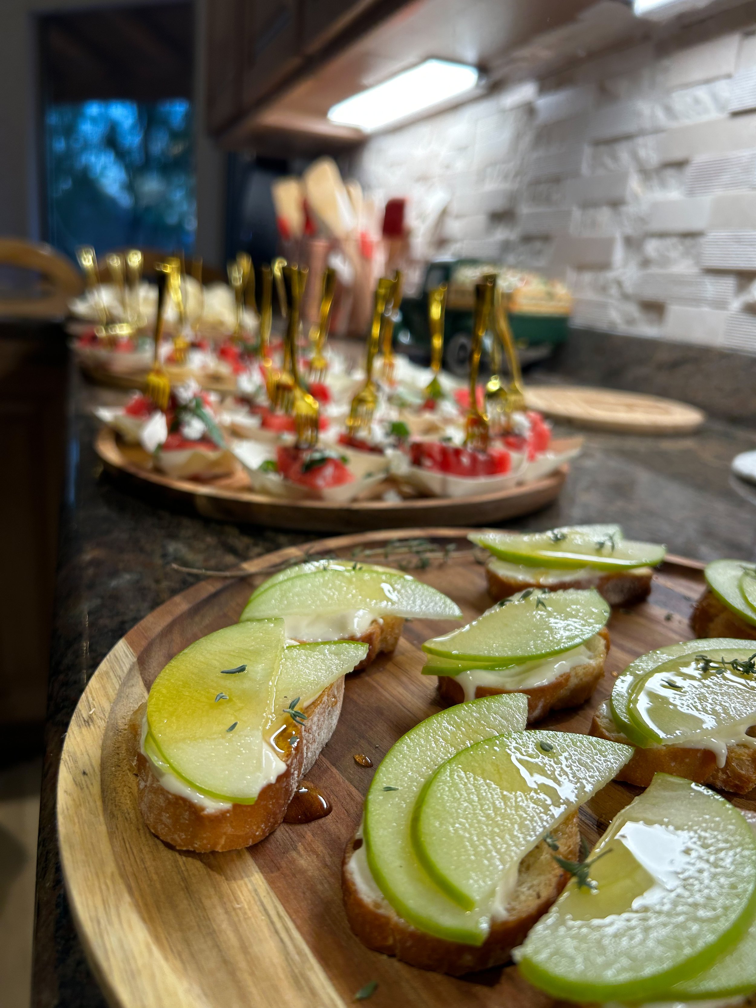 Close-up of crostini with sliced green apples, cream, and herbs on a wooden platter, with a blurred background of small appetizers with skewers and a kitchen counter.