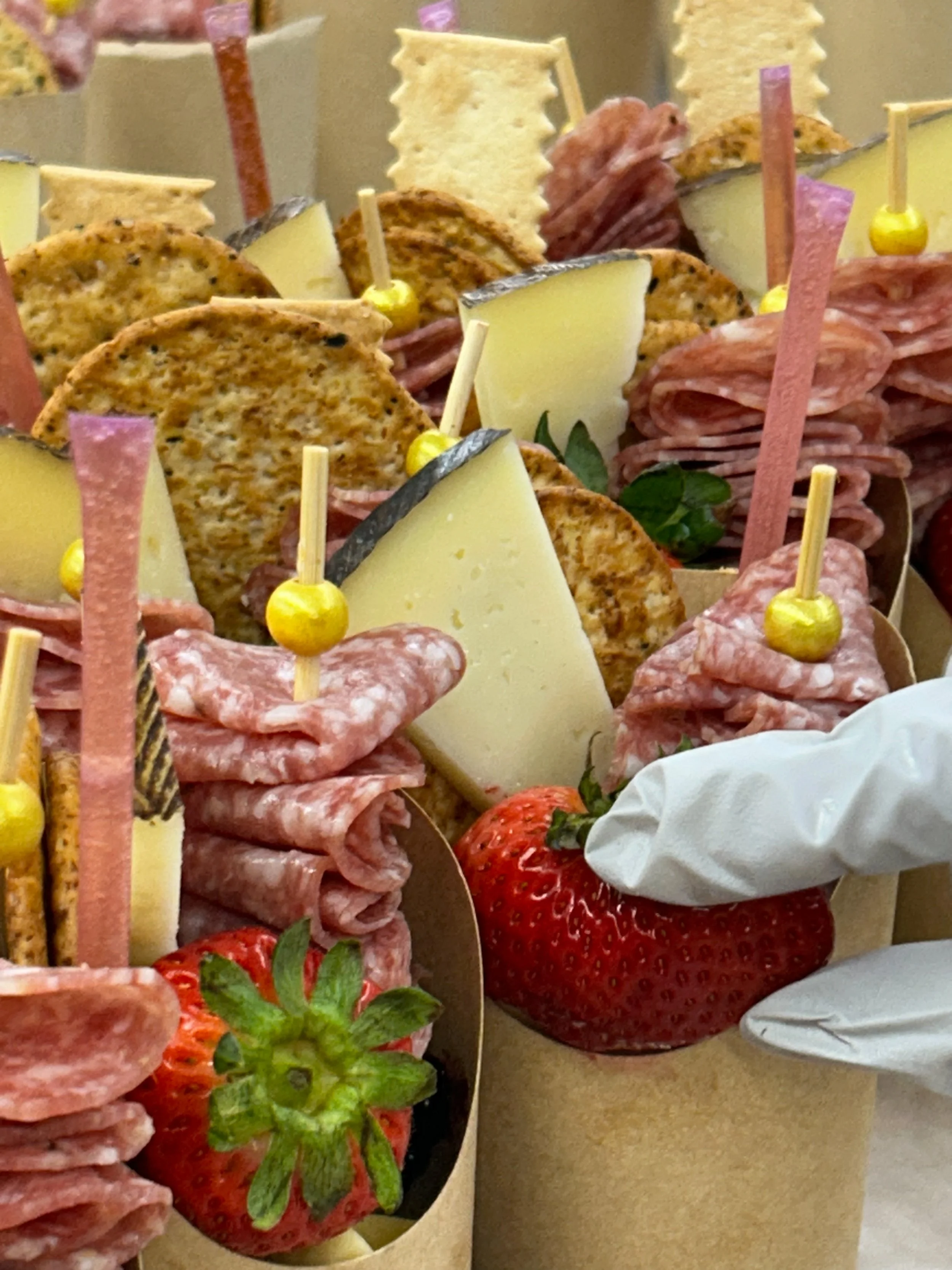 Assorted charcuterie and cheese appetizers with strawberries, crackers, cured meats, and cheese slices in small cups with skewers.
