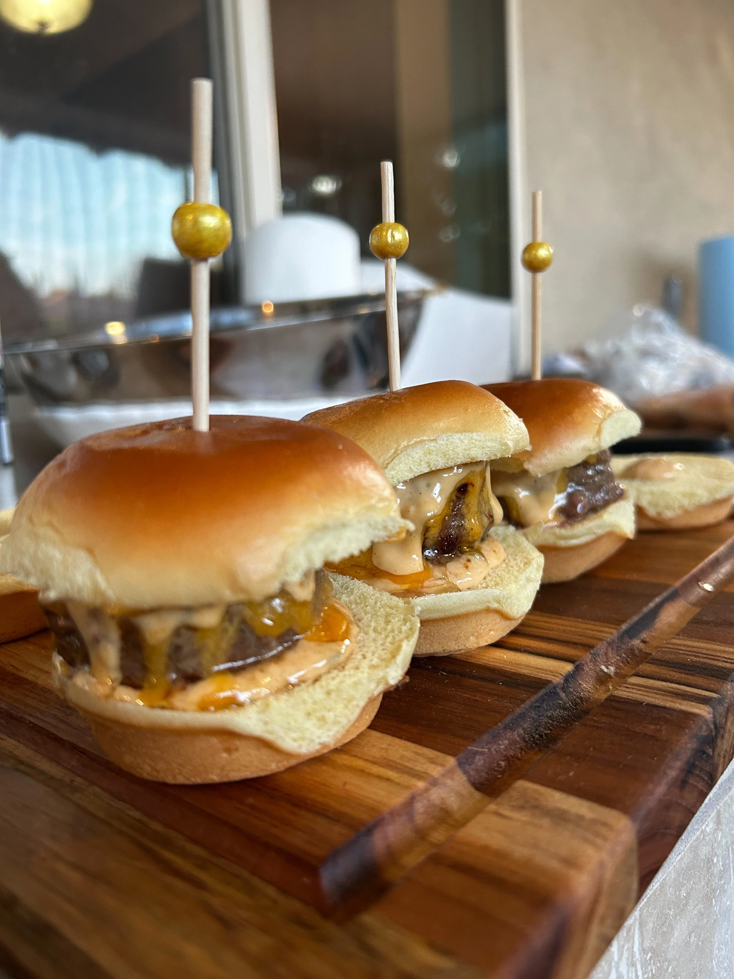Three mini sliders with beef patties, cheese, and sauces on a wooden serving board, each topped with a decorative toothpick with a small round ornament.