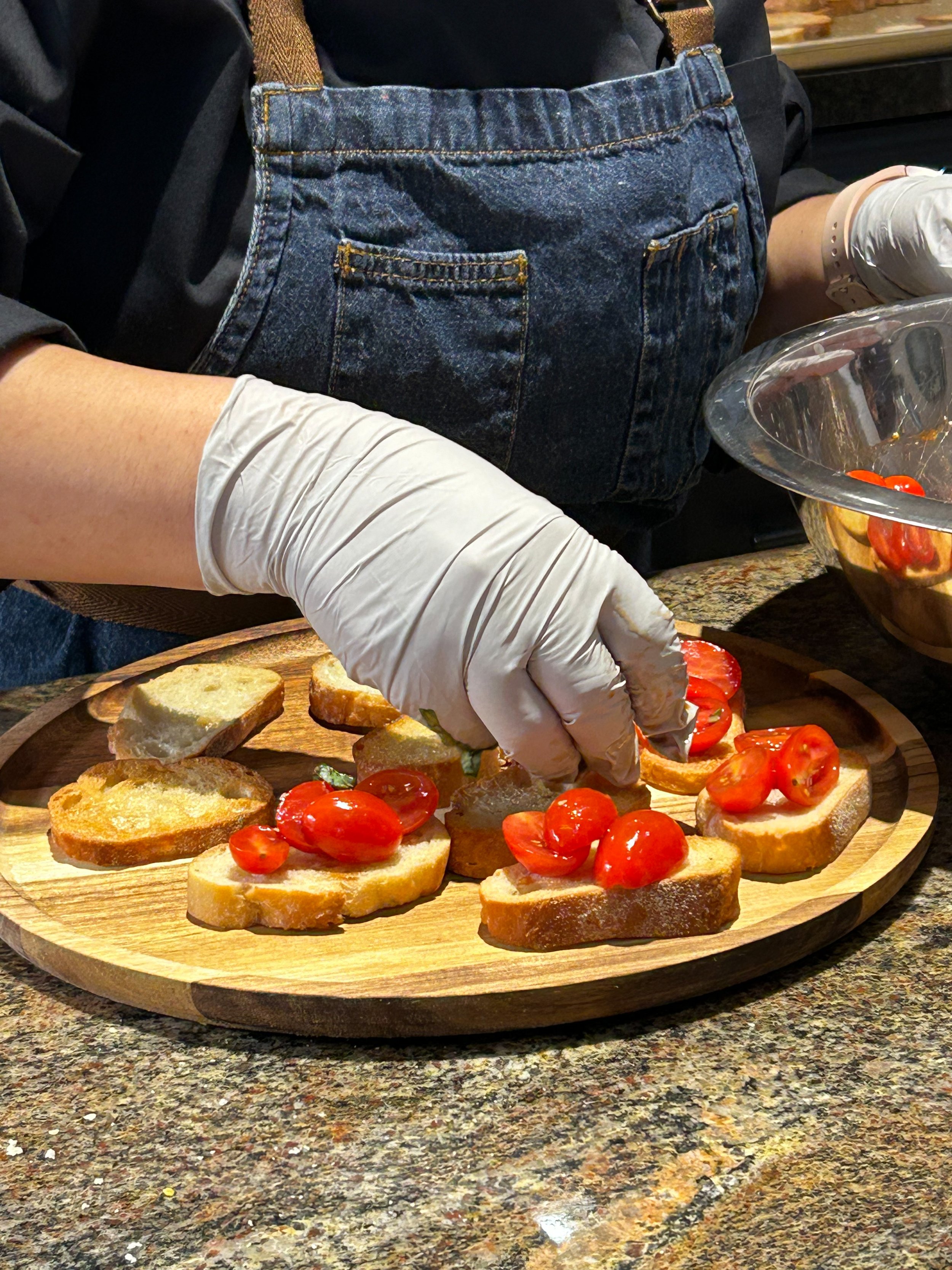 A person wearing gloves and a denim apron preparing bruschetta toppings on a wooden board, with halved cherry tomatoes. The person is assembling the dishes on a granite countertop.