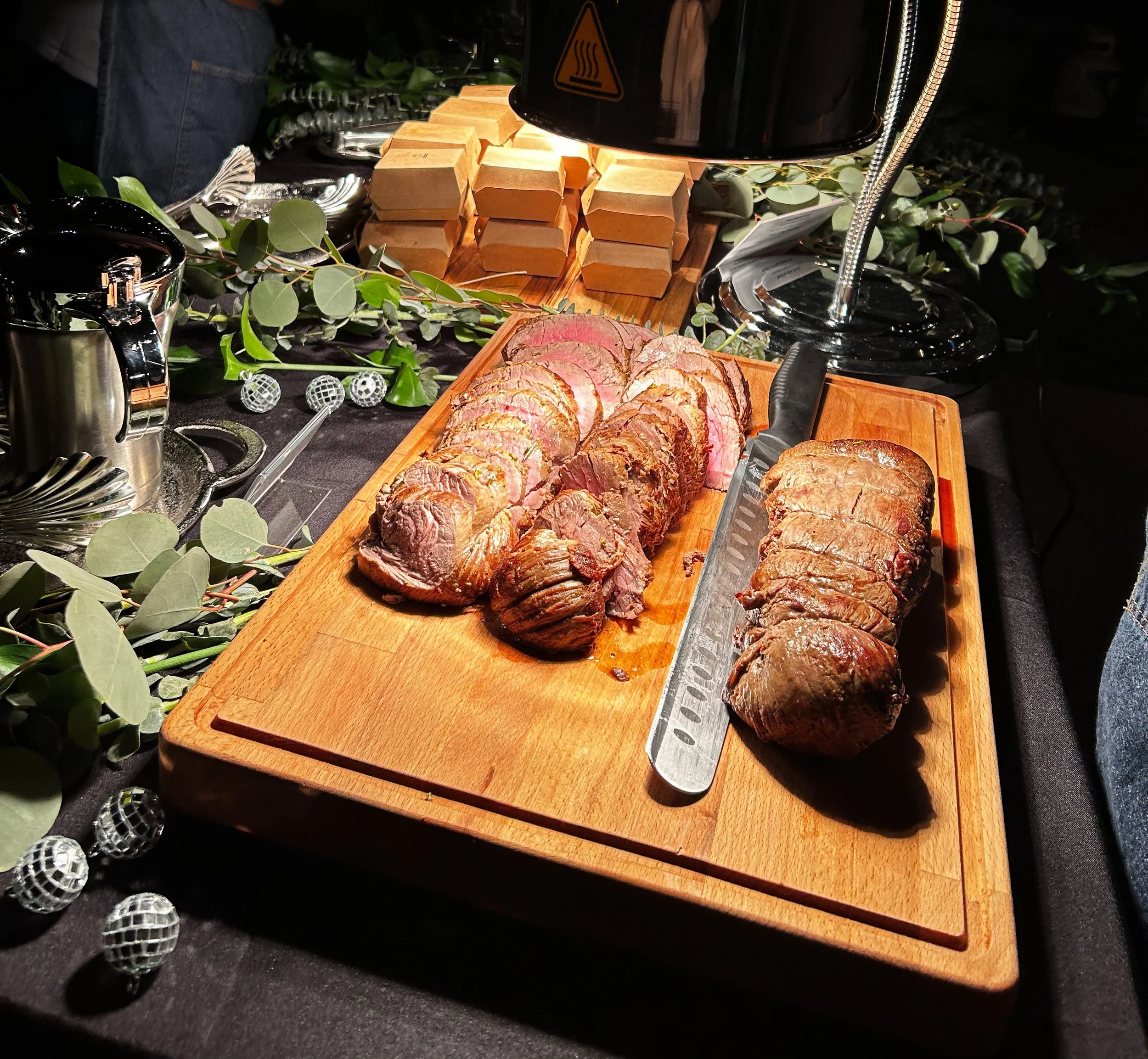 Sliced cooked beef tenderloin and roast beef on a wooden cutting board, illuminated by a lamp, with decorative greenery and small square bread rolls in the background.