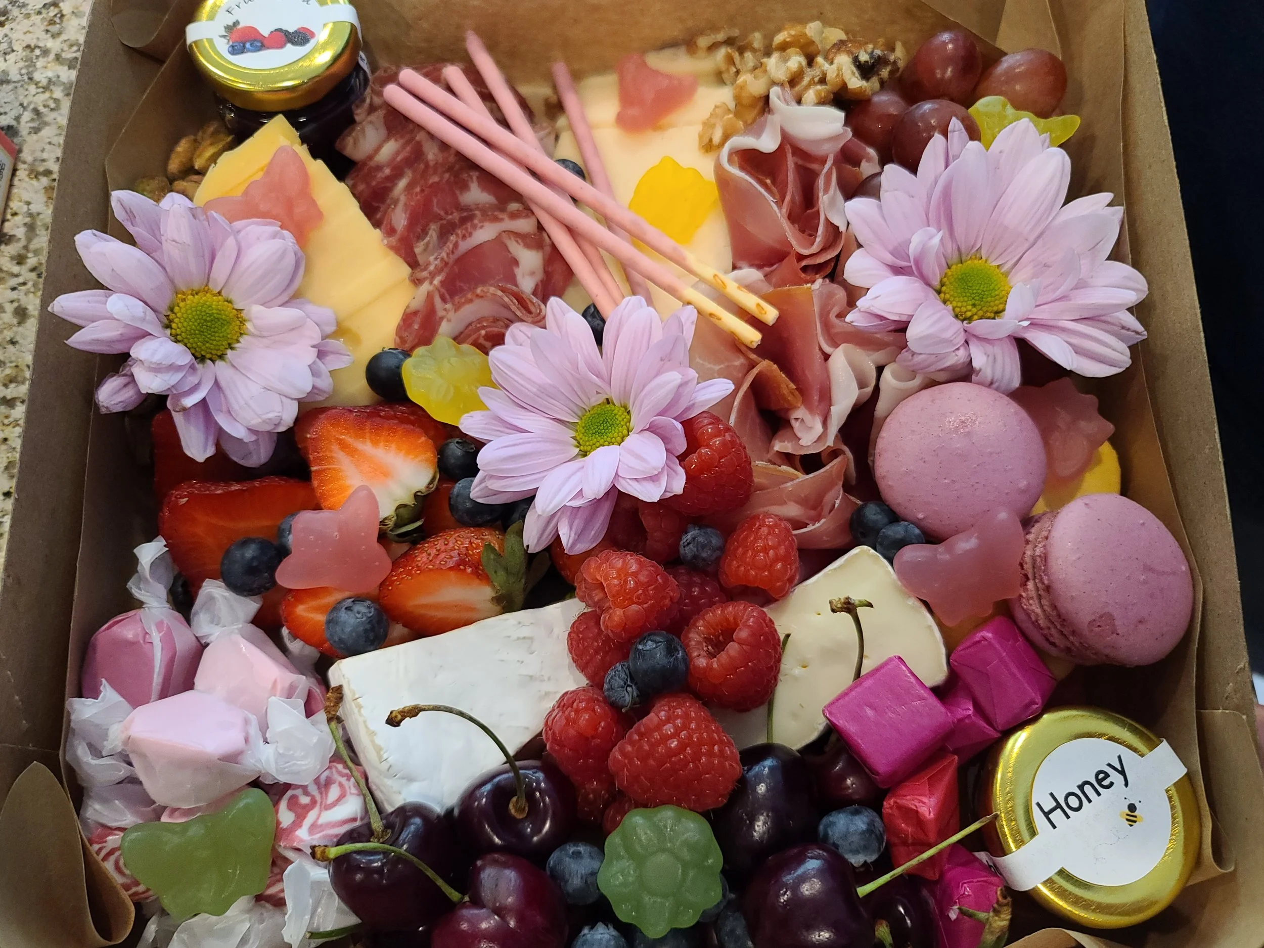 A gift box filled with strawberries, raspberries, blueberries, cherries, heart-shaped candies, pink macarons, cheese, ham, bread, honey, flowers, and colorful candies.