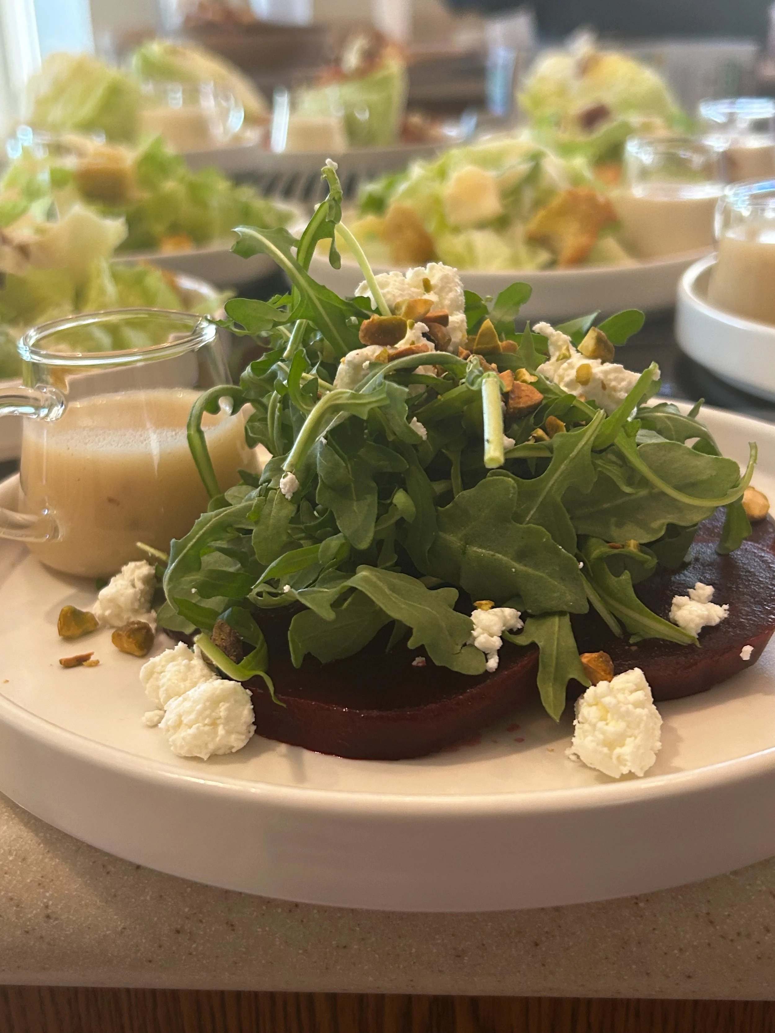 A plate of roasted beets topped with arugula, crumbled cheese, and chopped nuts, served with a side of salad and dressing.