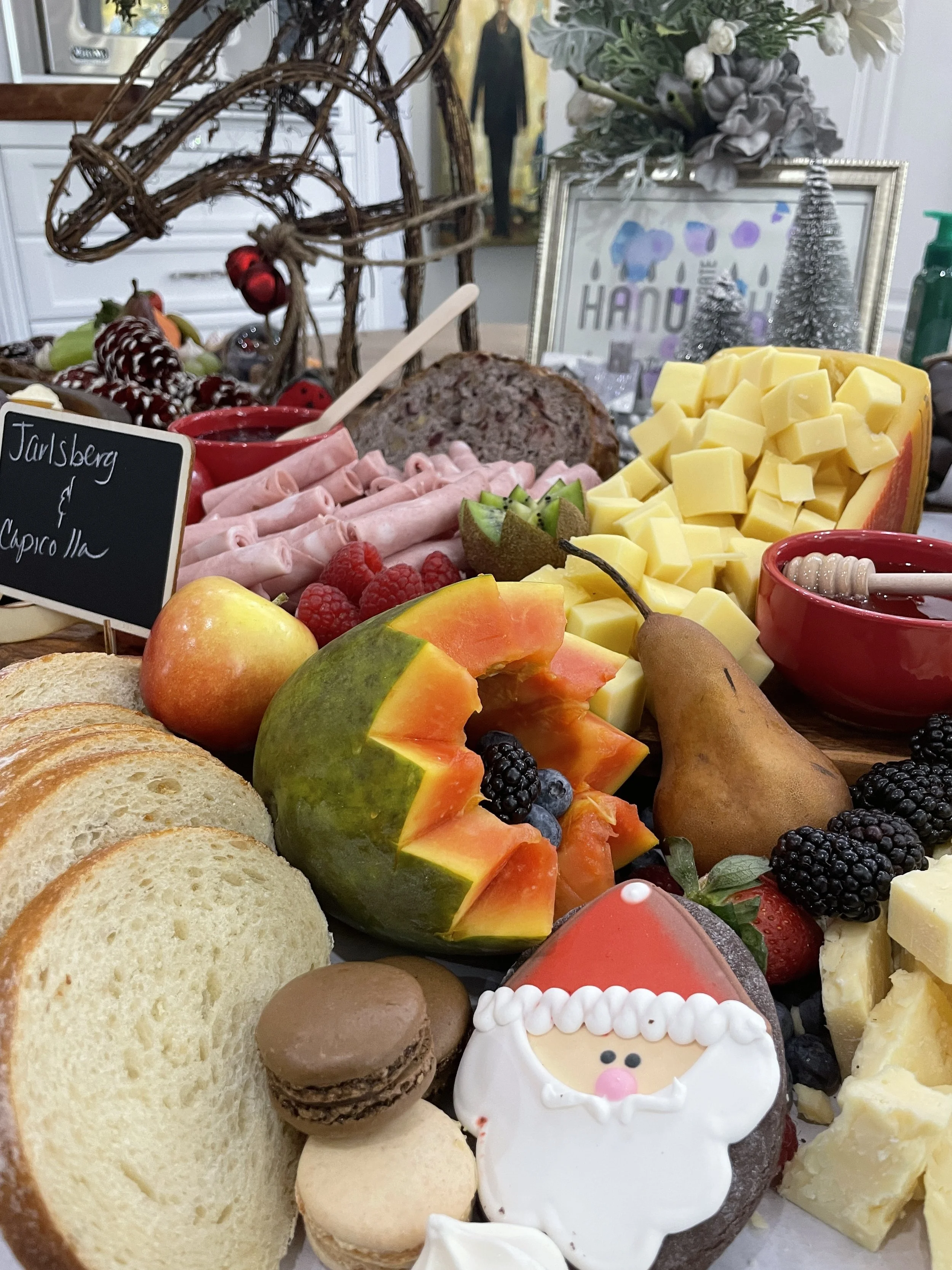 A festive holiday breakfast spread with cheese, fruits, bread, and desserts, including a Santa-shaped cookie and an apple-shaped cookie, surrounded by berries, sliced bread, cheese cubes, sliced ham, and decorative elements like small Christmas trees