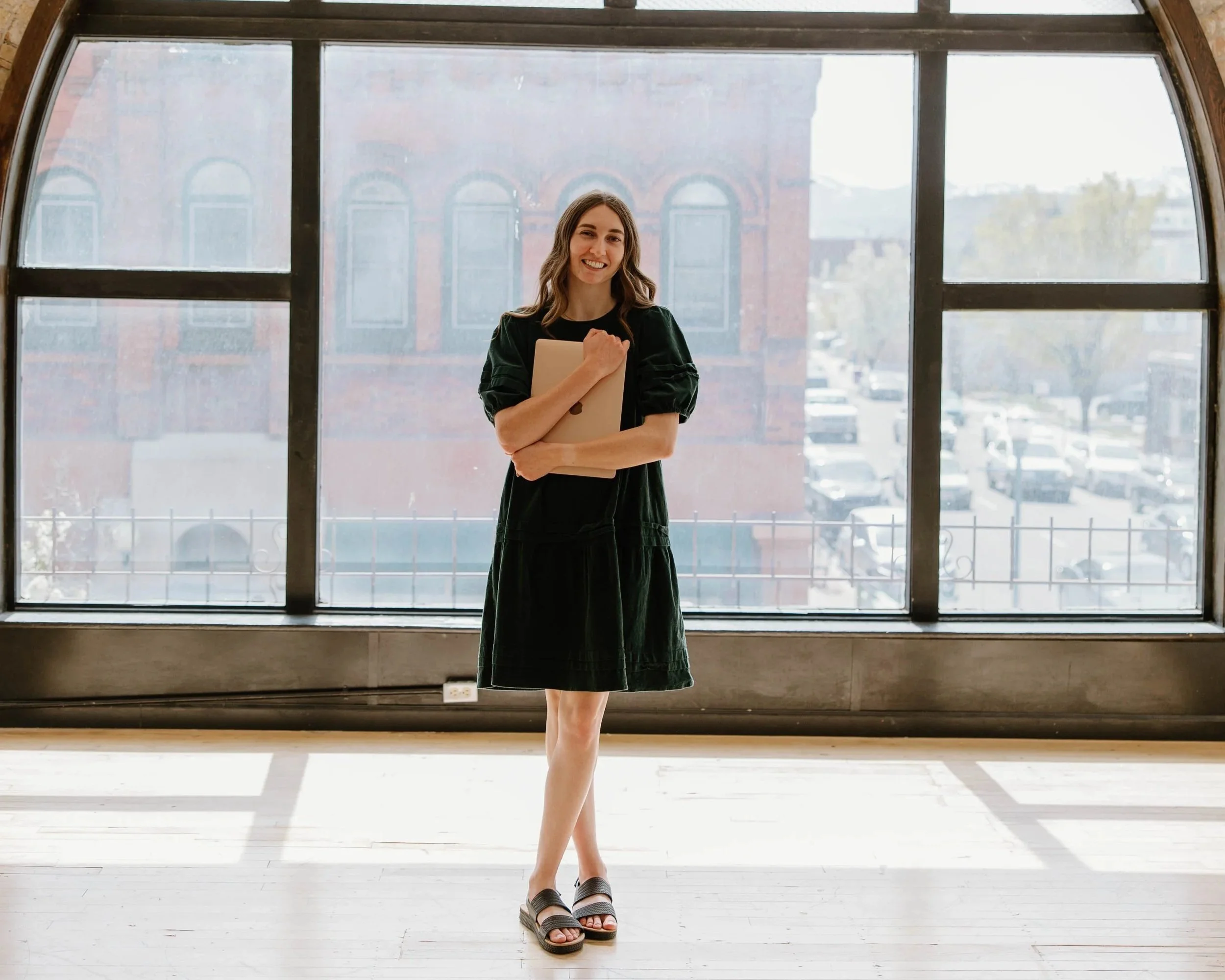 A young woman standing in front of a large window, smiling, holding a folder, wearing a black dress and sandals.