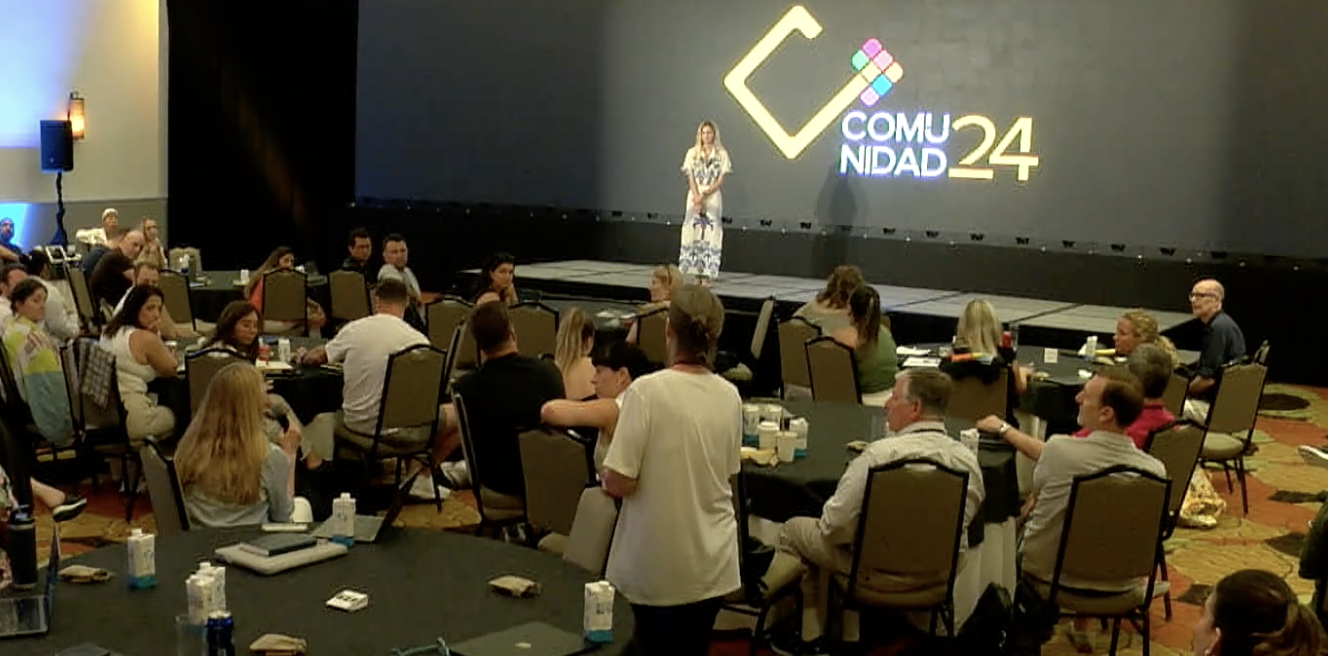 People attending a conference or event in a large room, seated at round tables with black tablecloths, facing a stage with a large screen displaying the logo "COMUNIDAD 24" and a woman speaking at the front.