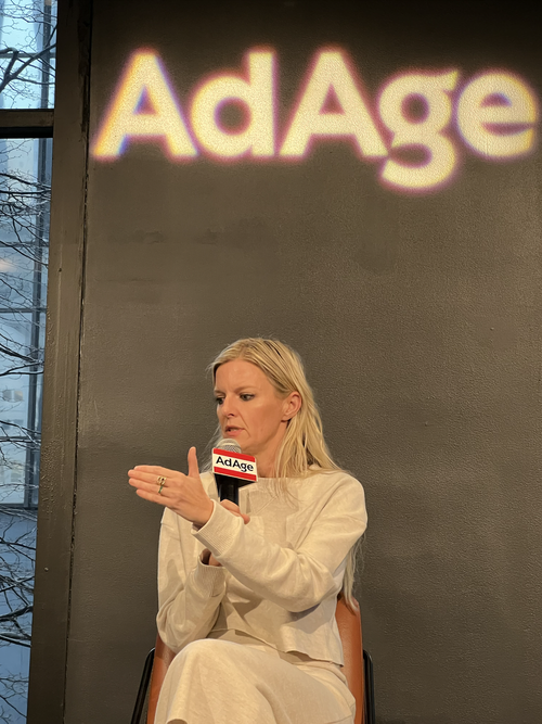 A woman sitting and speaking into a microphone labeled 'AdAge' during an event or interview, with a blurry logo projector displaying 'AdAge' on the wall behind her.