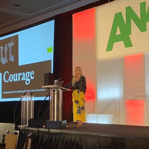 A woman standing at a podium on a stage with a large screen behind her displaying the word "Courage" and the logo of an organization.