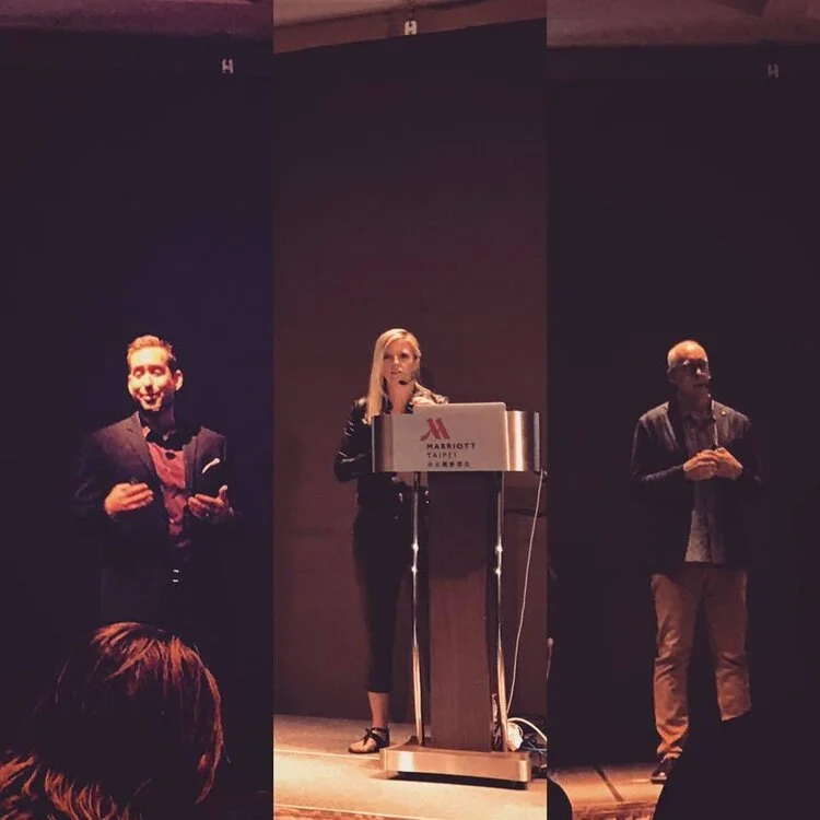 Three speakers presenting at a conference in a dark room with a large blank screen behind them. The woman in the center is standing at a podium with a Marriott hotel logo. The man on the left is smiling and speaking, and the man on the right has his