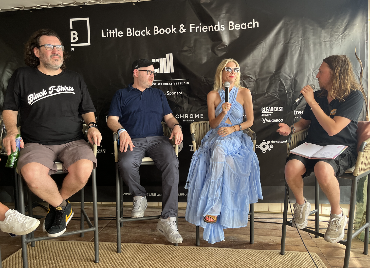 Four people seated on stools participating in a panel discussion, two men, one woman and one person with a microphone in their hand, backdrop with event information, casual attire, and a book or notes in hand.