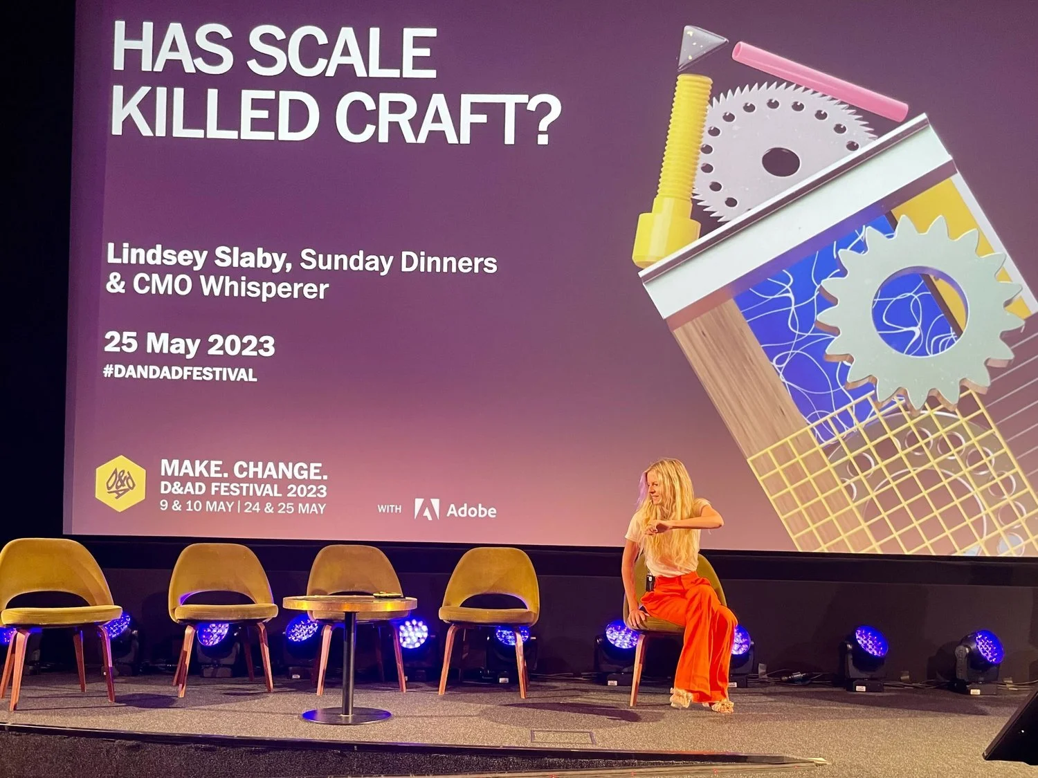 A woman with long blonde hair, wearing an orange skirt and white top, sitting on a chair on stage at a conference. Behind her, a large screen displays the text "Has Scale Killed Craft?" along with details of the event, date, speaker, and sponsors. Th
