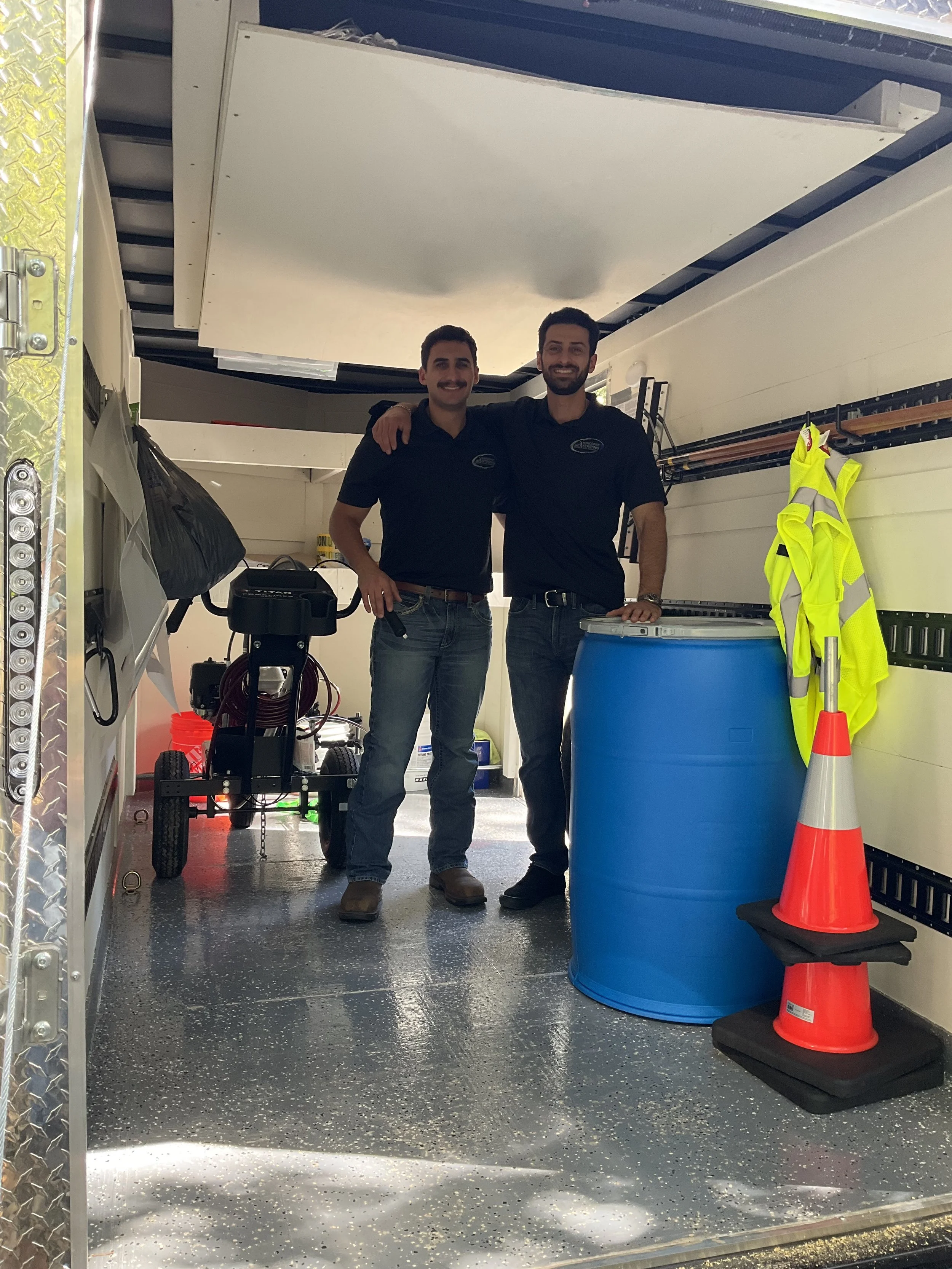 Two men smiling inside a service truck, wearing dark shirts, with safety equipment, a large blue barrel, and a cone nearby.