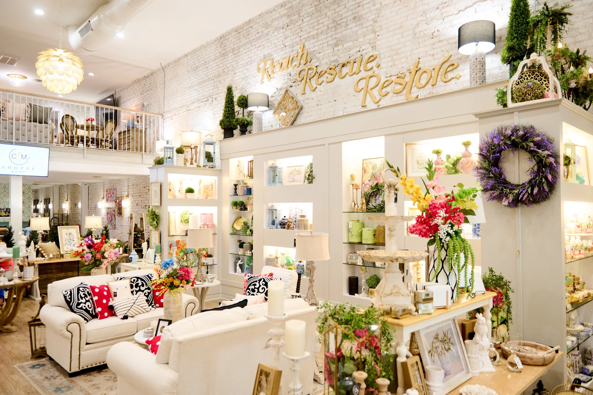 Interior of a boutique shop with white brick walls decorated with gold signs reading 'Ranch Rescue Restore'. The shop is filled with white furniture, floral arrangements, and decorative items on shelves and tables.