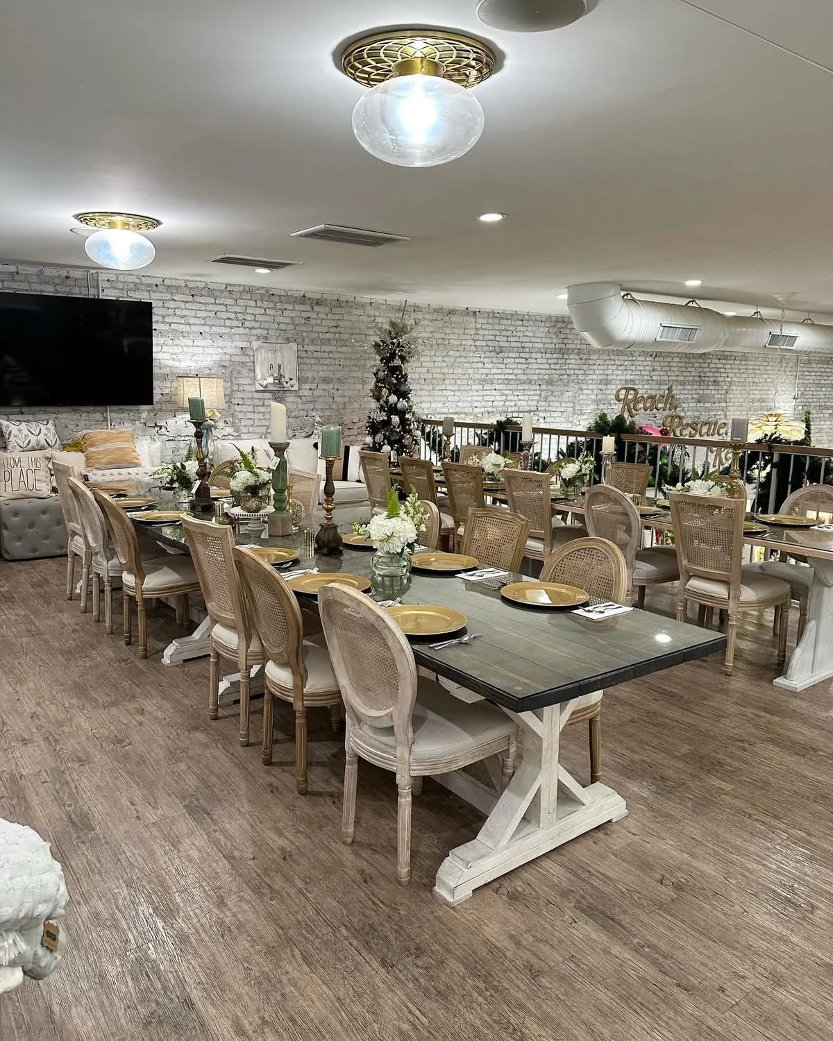 A decorated dining area with a long table set with gold plates, silverware, and floral centerpieces, with a Christmas tree and holiday decor in the background.