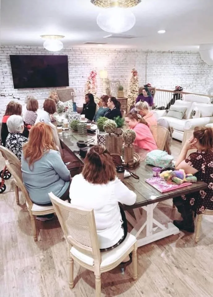 A group of women seated around a long dining table in a cozy room, with some decorated Christmas trees in the background, a large wall-mounted TV, and a white brick wall.