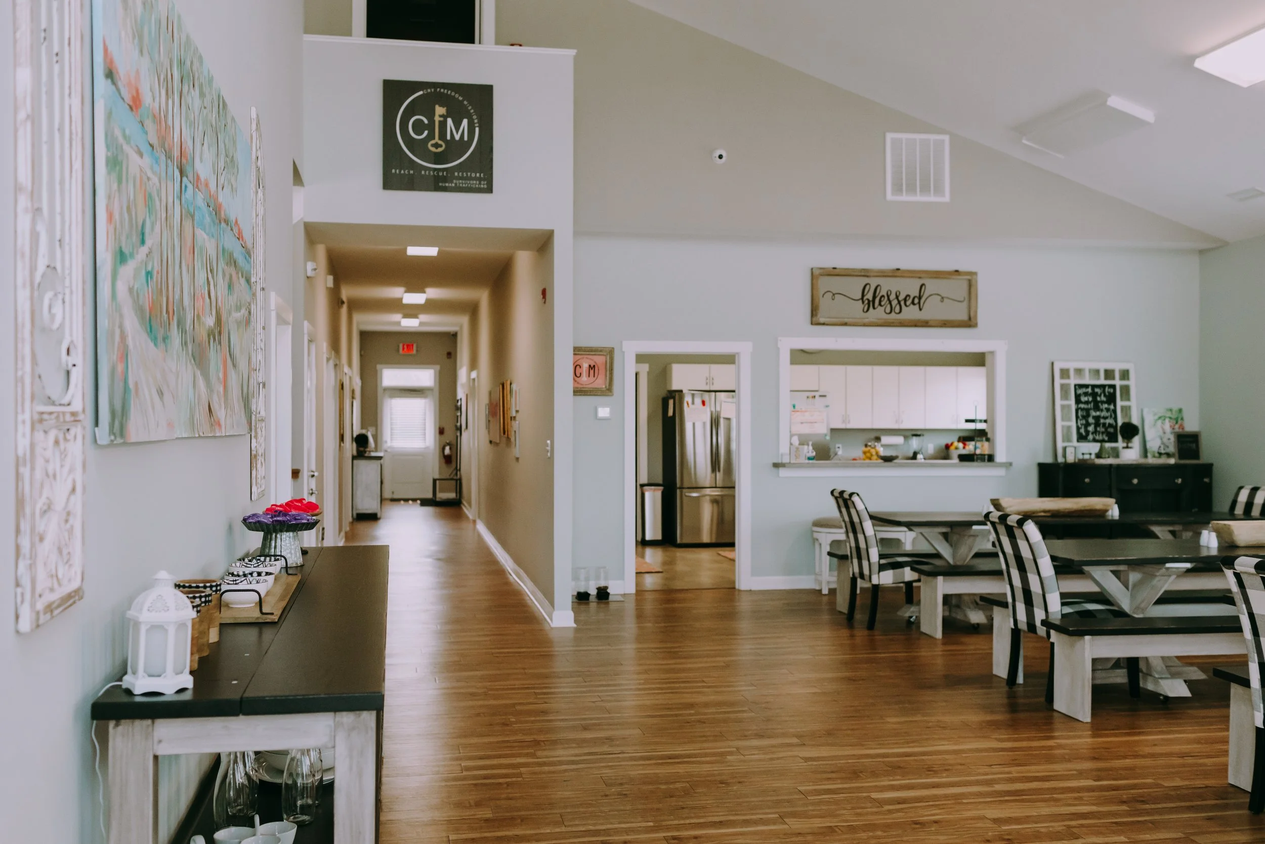 A cozy dining area with checkered black-and-white chairs, a wooden table, and decorative wall signs, with a view into the kitchen and a hallway leading to other rooms.