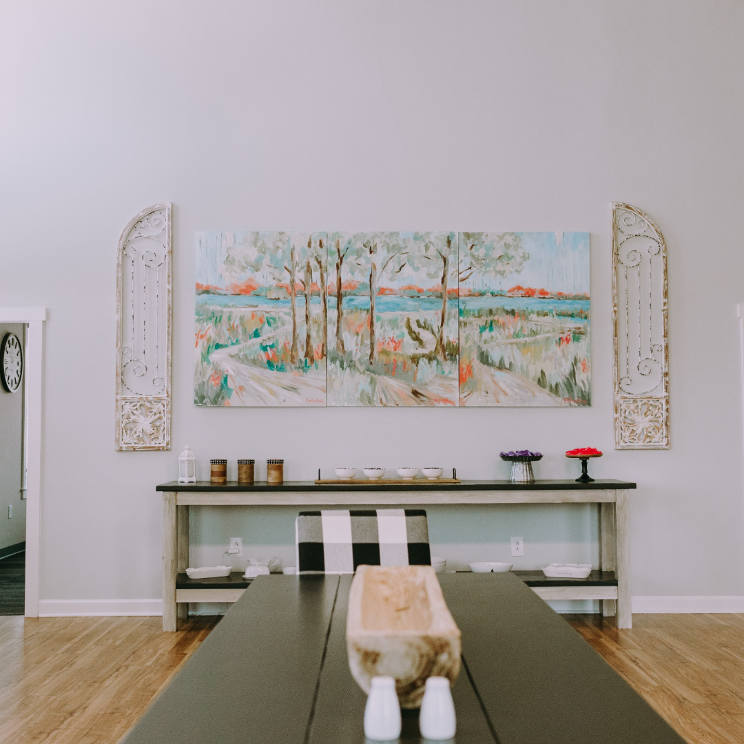 Dining room with a long black table, white and striped chairs, a wooden centerpiece, wall art of a three-panel landscape painting, white decorative wall panels, and a sideboard with bowls and decorative items.