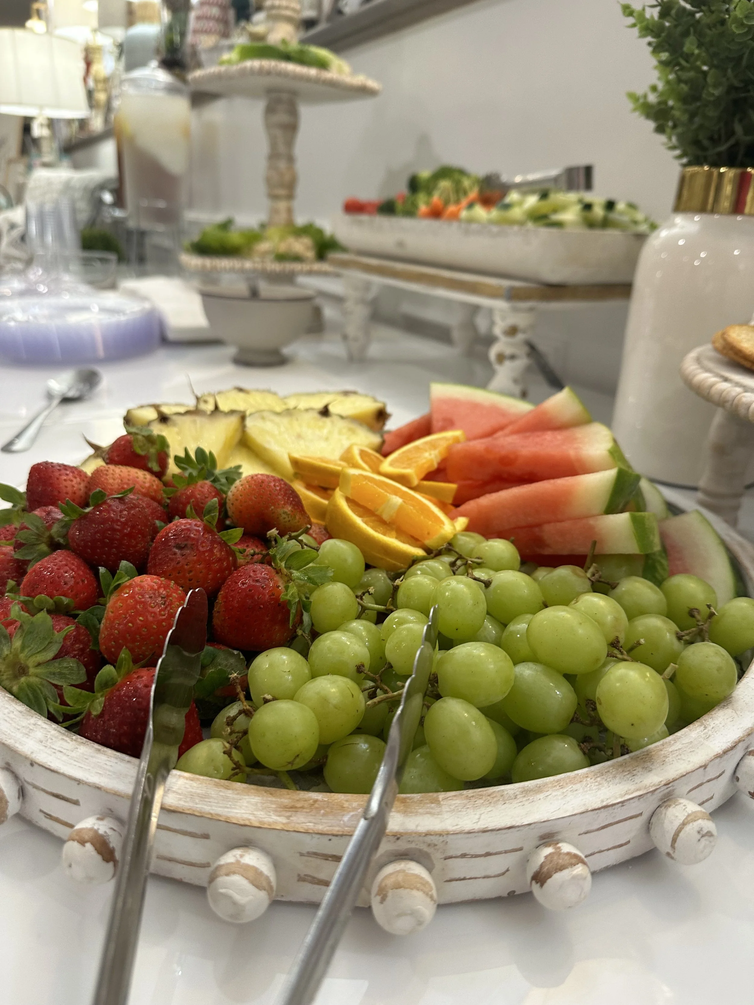 A white woven basket filled with strawberries, green grapes, sliced watermelon, pineapple, and orange slices, with tongs on the basket.