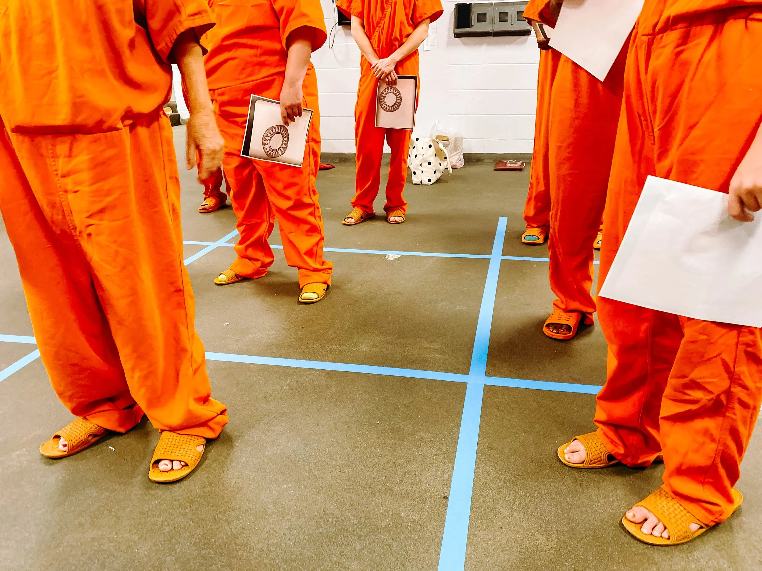 Several people dressed in orange prison uniforms standing in a room with blue tape on the floor.