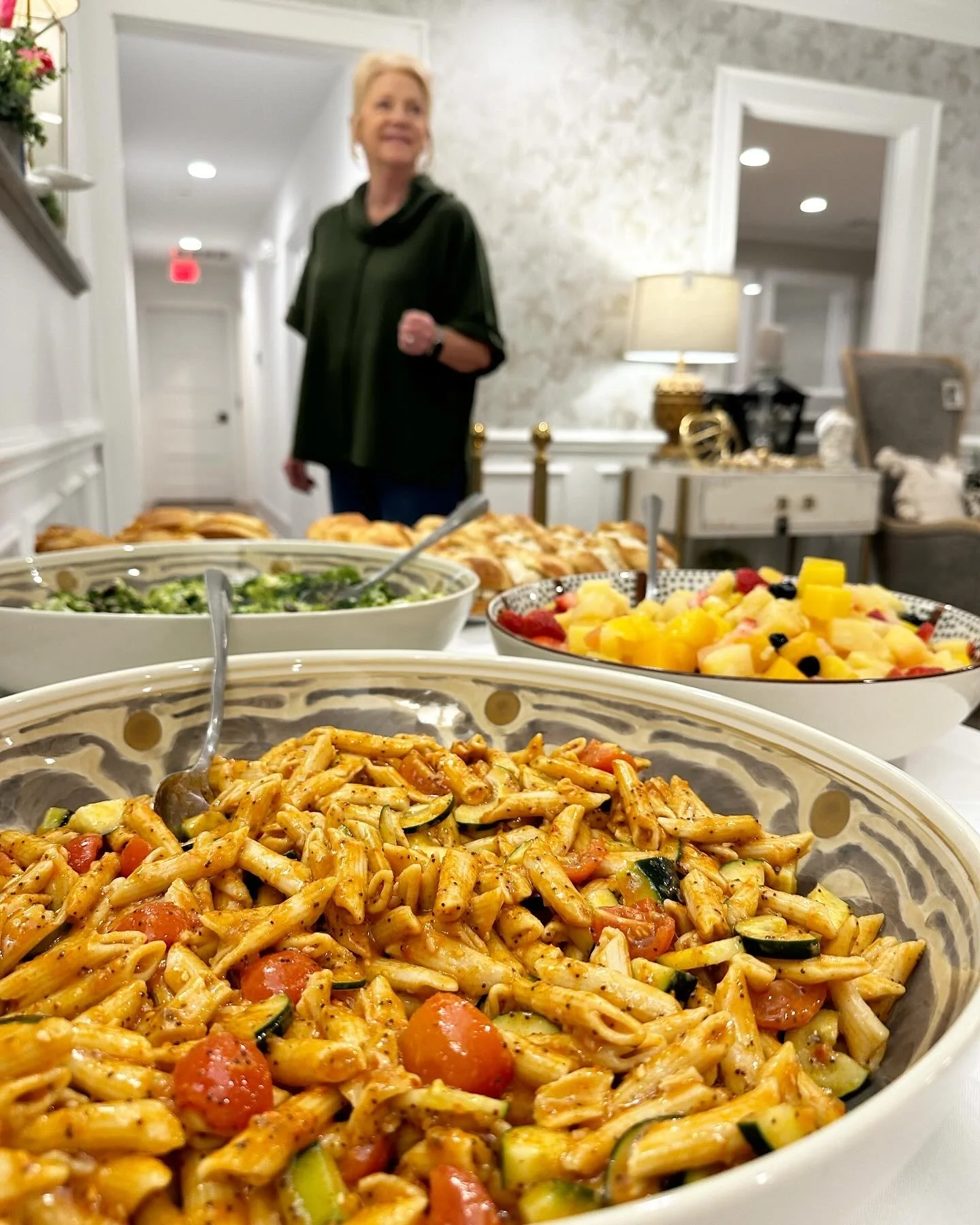 Bowl of pasta salad with cherry tomatoes and zucchini in the foreground, with other bowls of fruit salad and salad behind on a table, a woman in a green top smiling in a decorated indoor setting.
