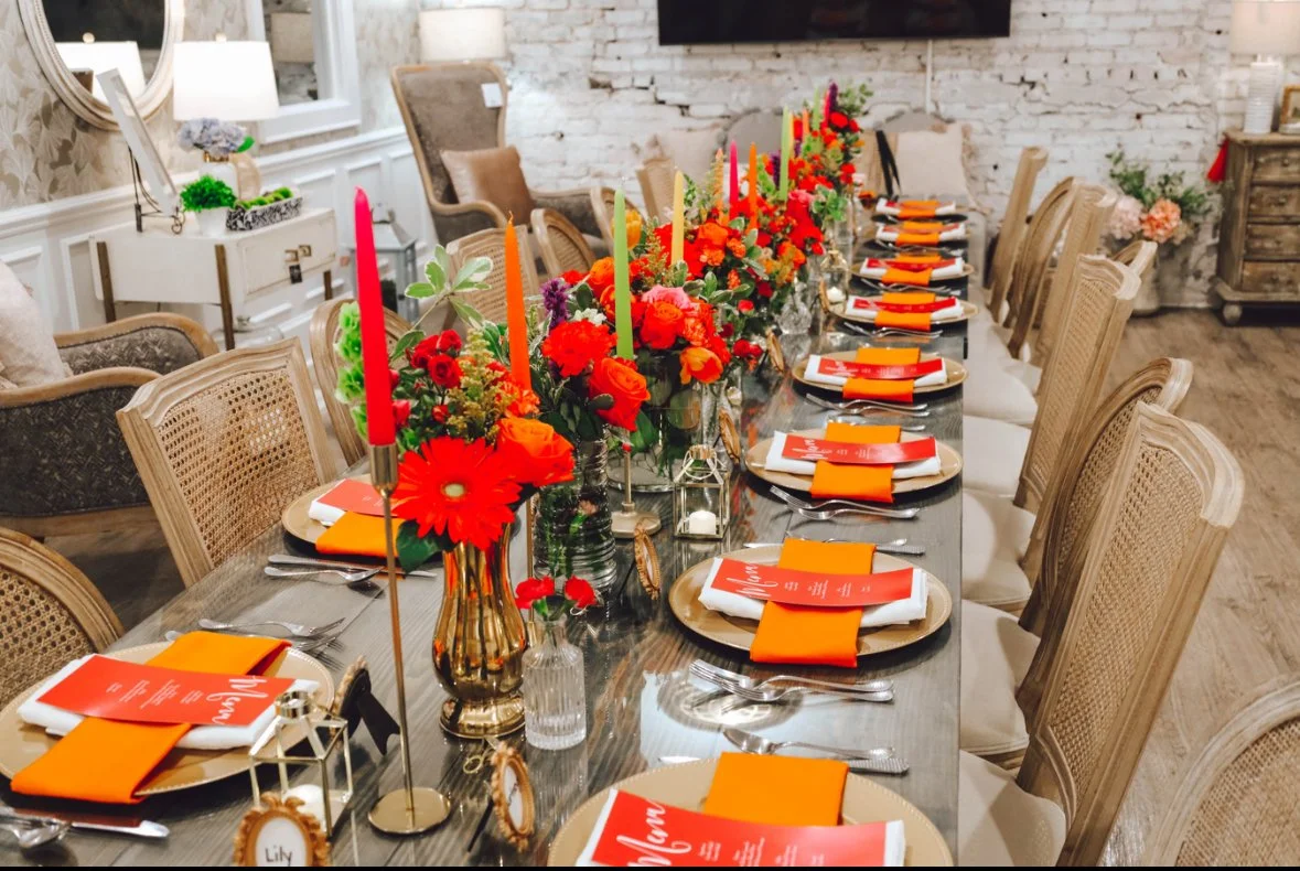 A long dining table set for a celebration, decorated with colorful floral arrangements and tall, colorful candles. Each place setting includes a plate, napkin, and printed menu. The room has a cozy, rustic style with white brick walls and elegant cha