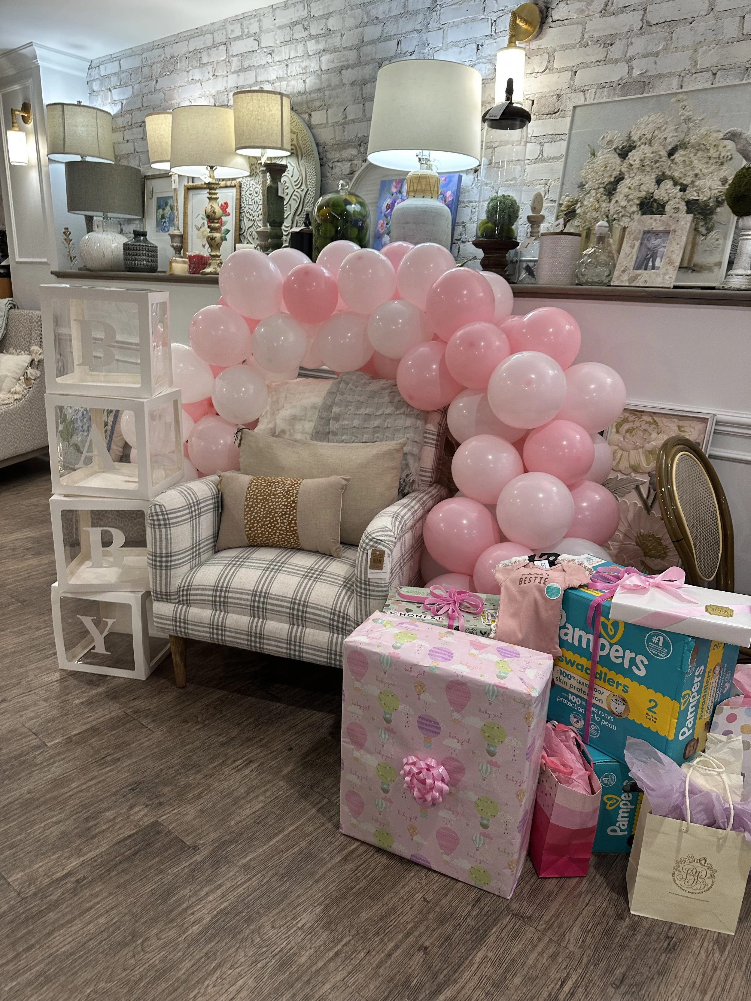 A decorated space for a baby shower with pink and white balloons, a plaid armchair with cushions, and presents including a gift box with a pink ribbon, all set against a white brick wall with lamps and framed photos.
