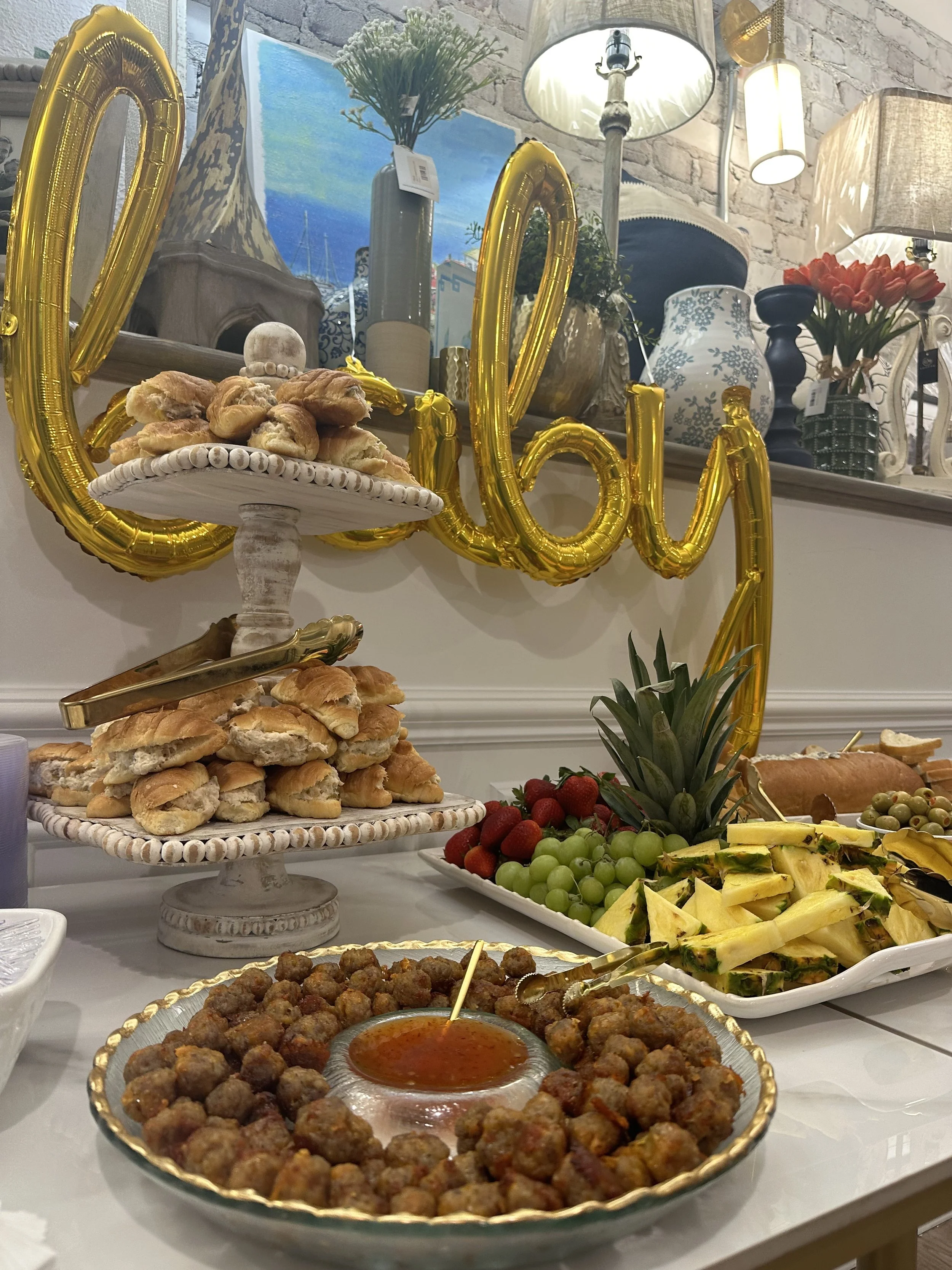 A table set with snacks including a large platter of meatballs with dipping sauce, sliced pineapple, grapes, strawberries, and pastries, with golden balloon letters spelling 'Love' behind the table. In the background, there are decorative vases, lamp