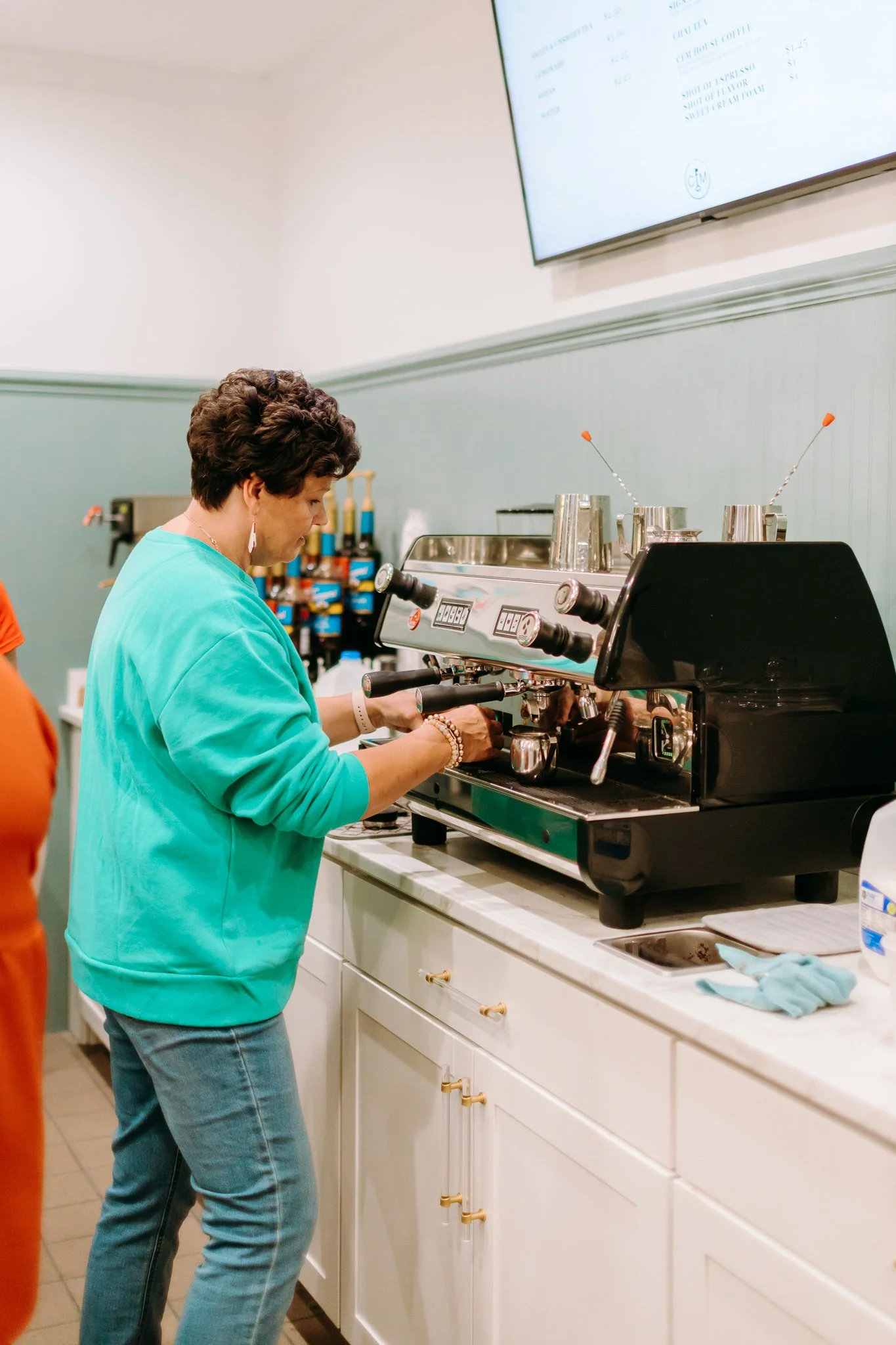 A woman in a turquoise sweatshirt making espresso using an espresso machine in a coffee shop.