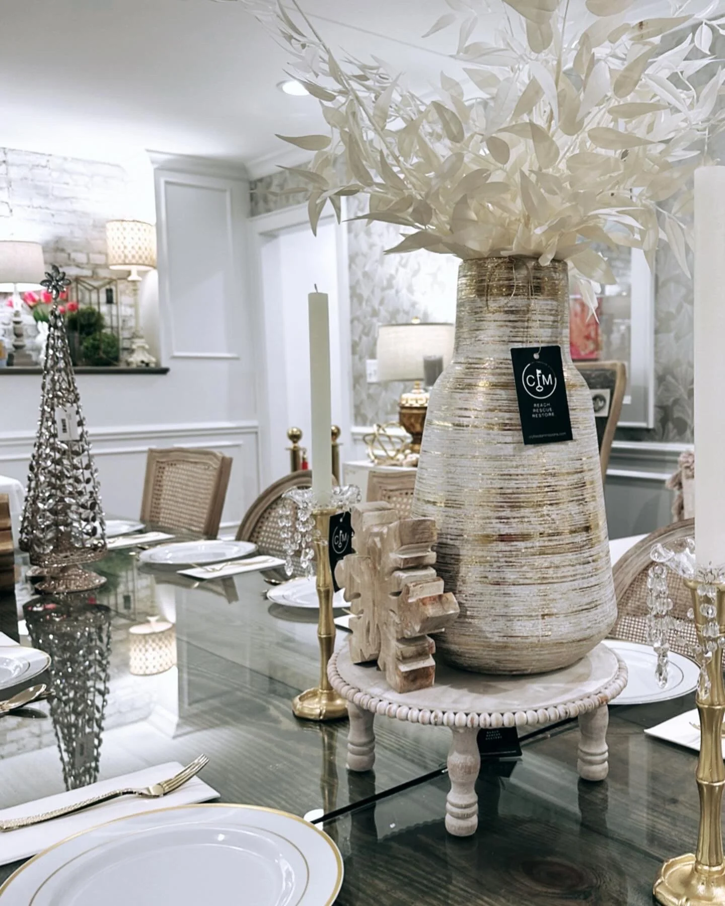 Decorative holiday table centerpiece featuring a large rustic vase with white leaves, surrounded by candles, ornaments, and dinnerware in a well-decorated dining room.