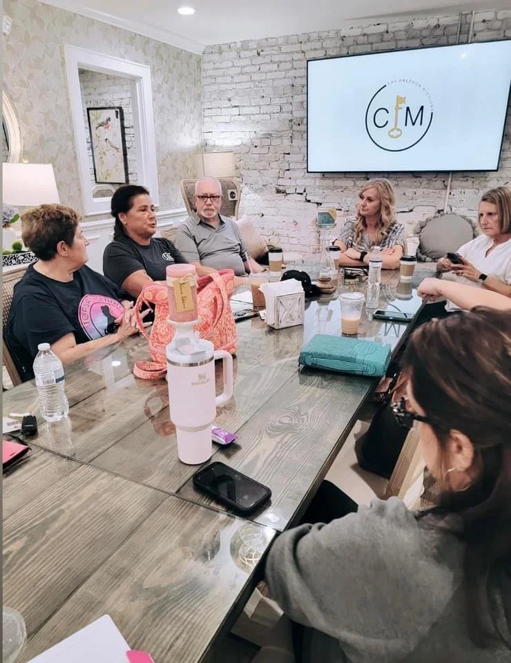 A group of seven people sitting around a large wooden conference table in a room with light-colored walls, some brick sections, and a large TV screen displaying a logo with the initials 'C M'. The table has water bottles, coffee drinks, a pink water