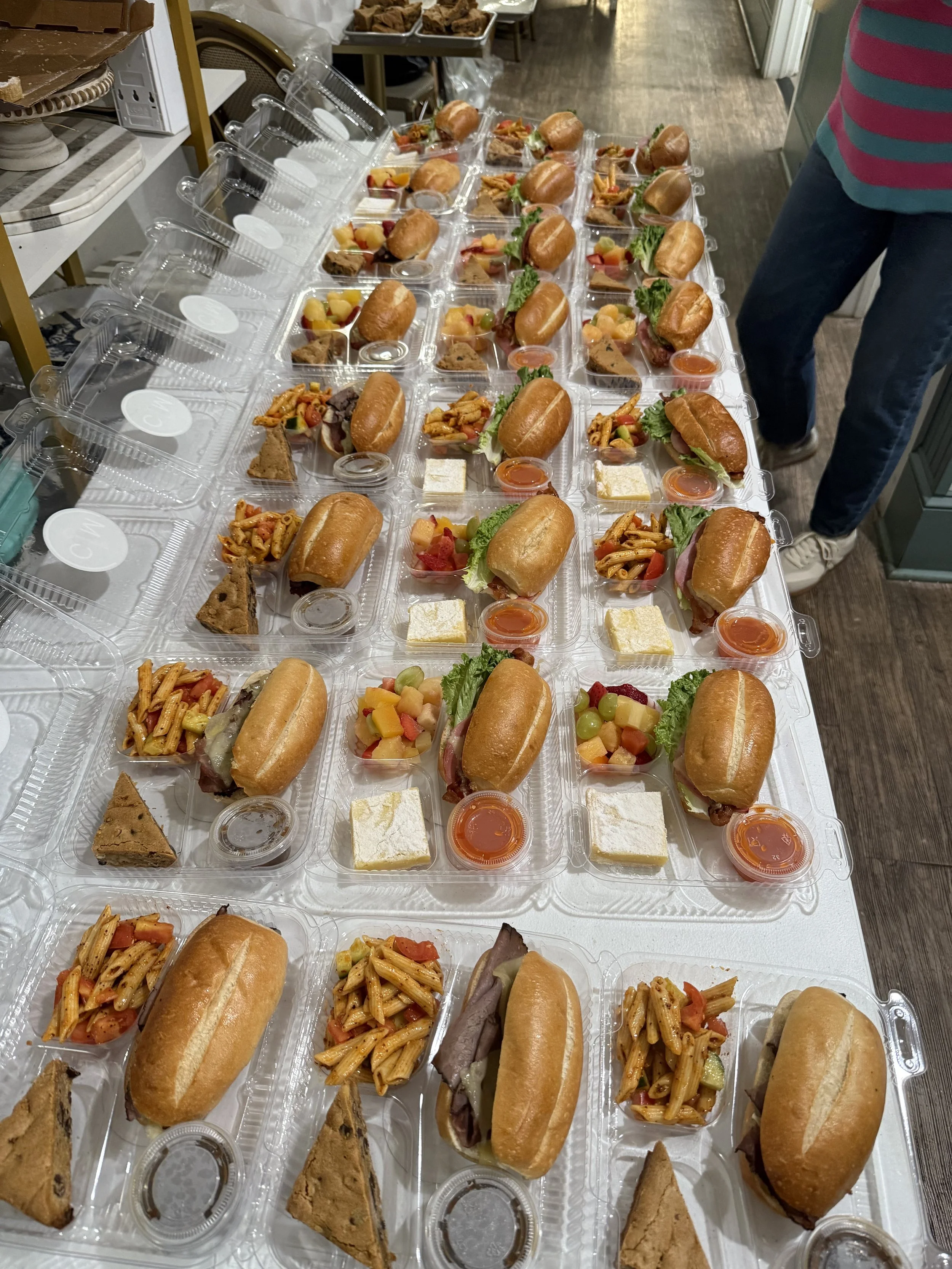 Pre-packaged meals arranged in clear plastic containers on a table, each containing a sandwich, a side of pasta salad, a small slice of cake, a piece of fruit, and a small cup of sauce or dip.