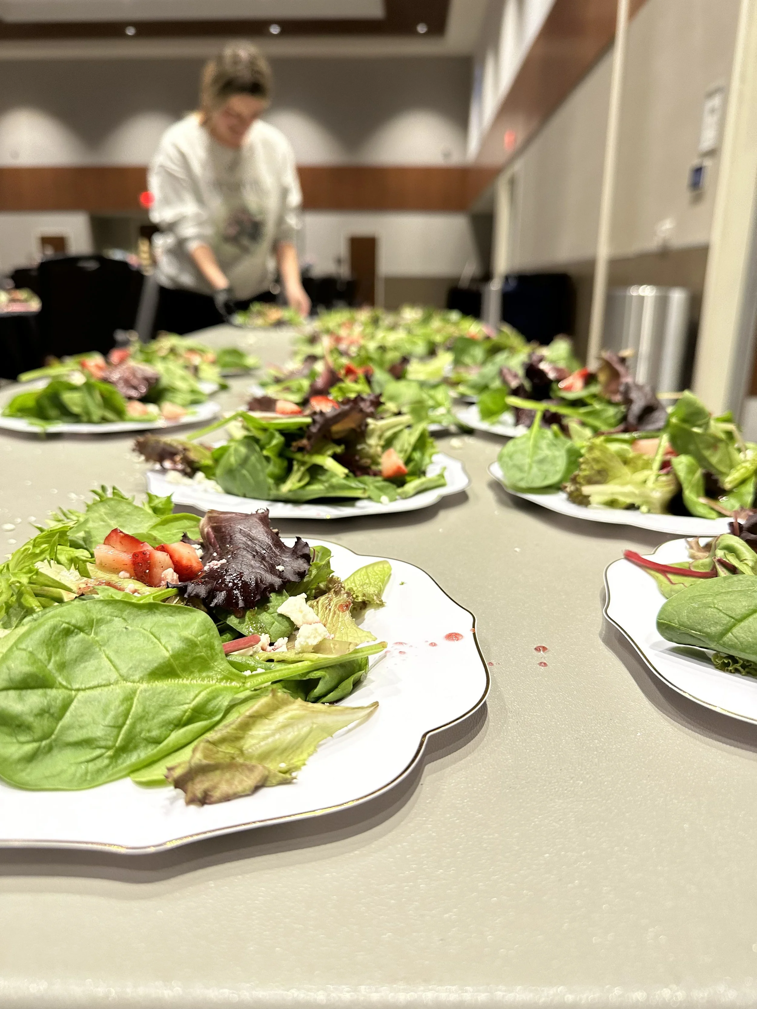 Several plates of mixed green salads with strawberries on a table, with a woman in the background preparing more salads in a conference or meeting room.
