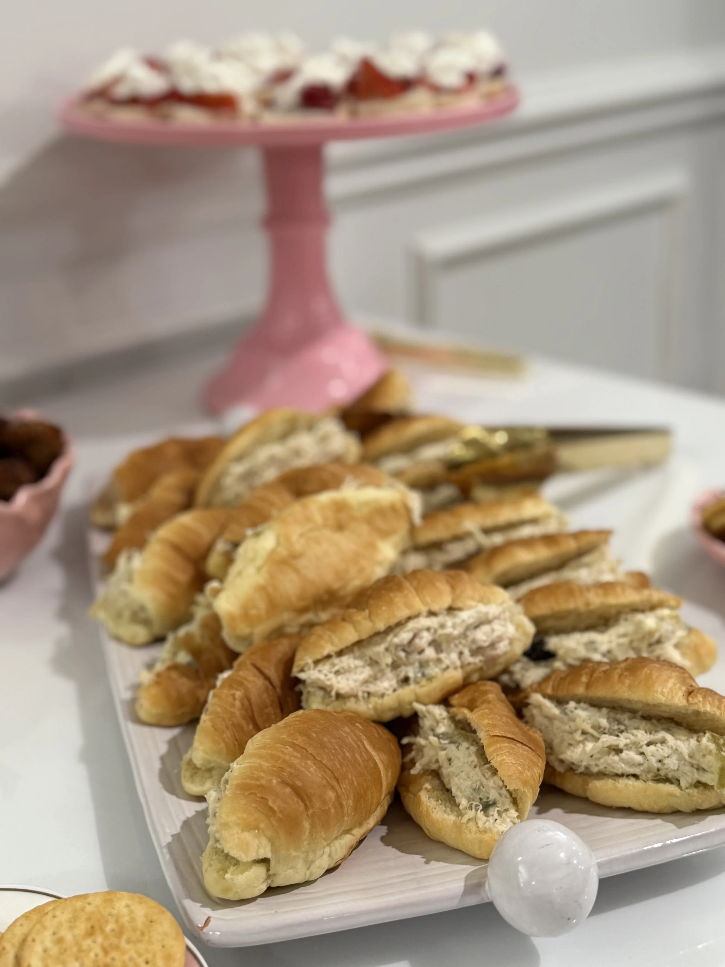 A white platter filled with assorted cookies and pastries on a table with a pink cake stand holding cupcakes in the background.