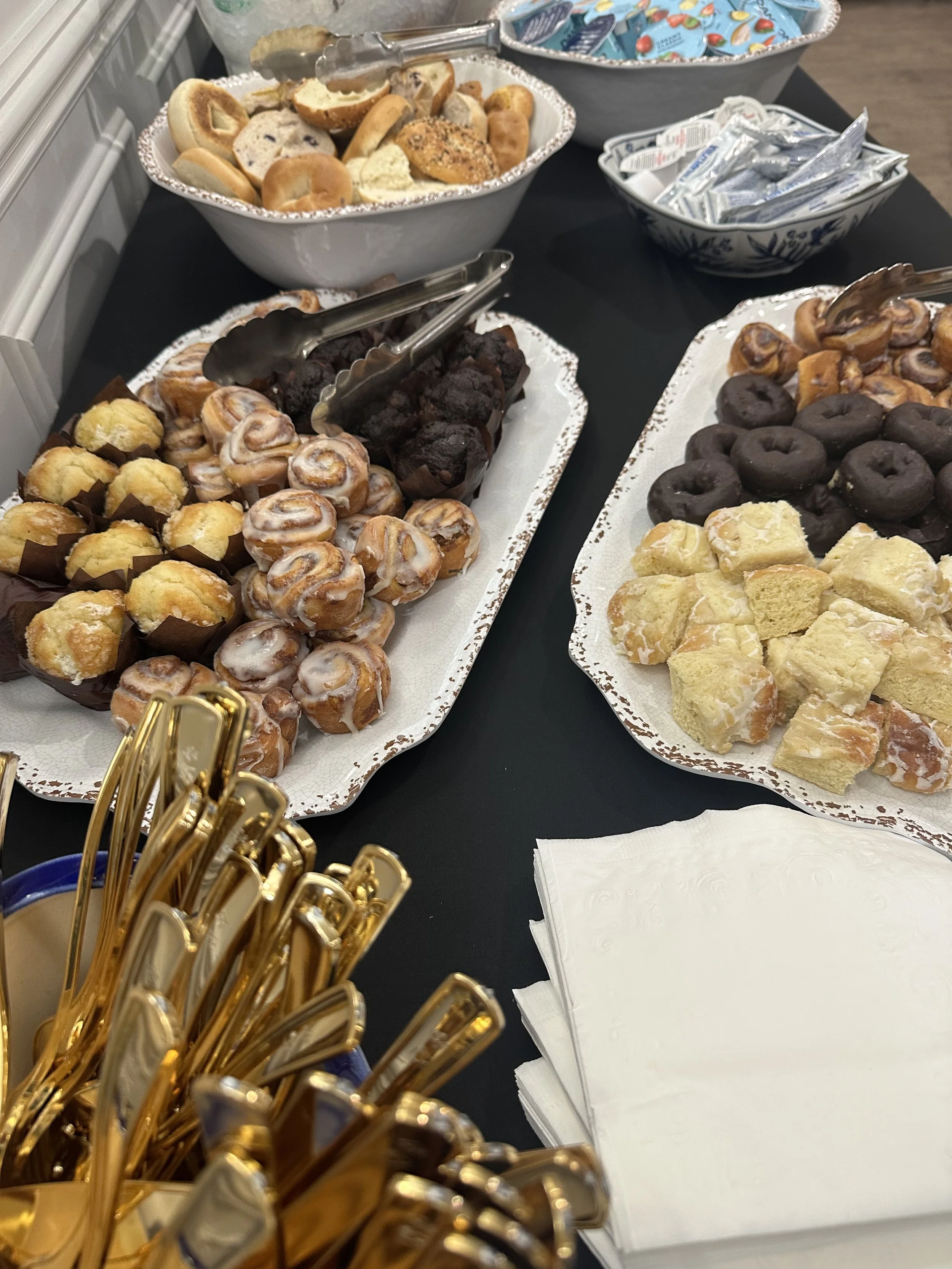 Assorted cookies and donuts on decorative trays at a dessert table, with napkins and gold-colored utensils nearby.