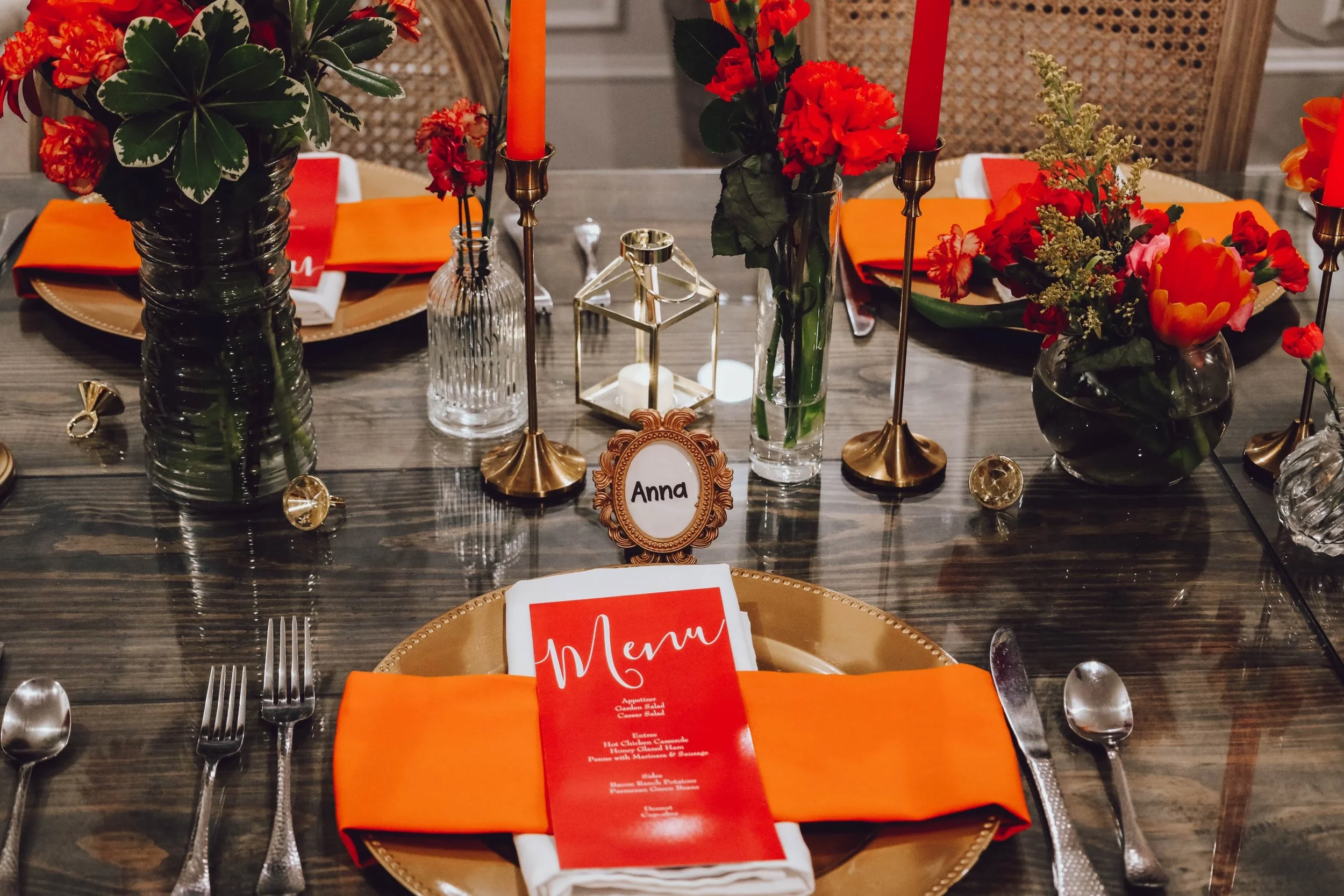 A formal dining table set with floral centerpieces, gold chargers, orange napkins, silverware, a menu card, and a name tag with 'Anna', decorated with red and orange flowers and candles.