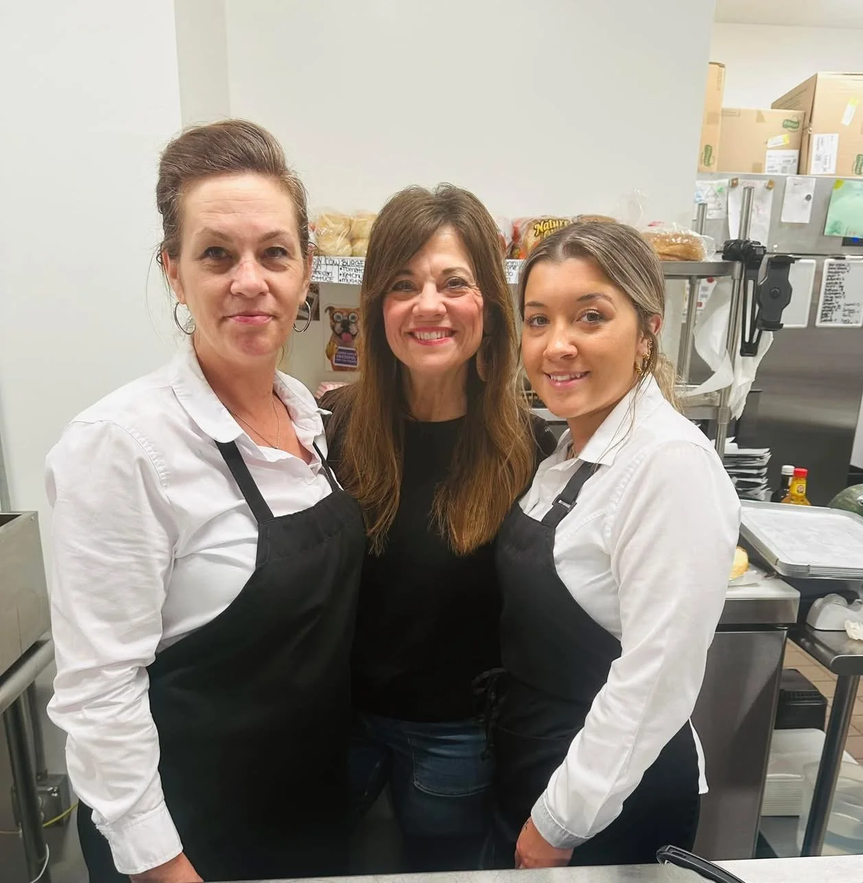 Three women in a kitchen, two wearing white shirts and black aprons, and one in a black shirt, all smiling at the camera.