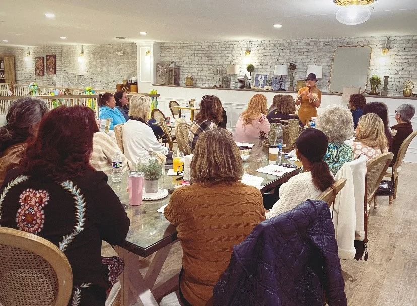 A woman in a hat giving a presentation to a group of women seated at a large table in a cozy, well-lit room with white brick walls and decorative items.