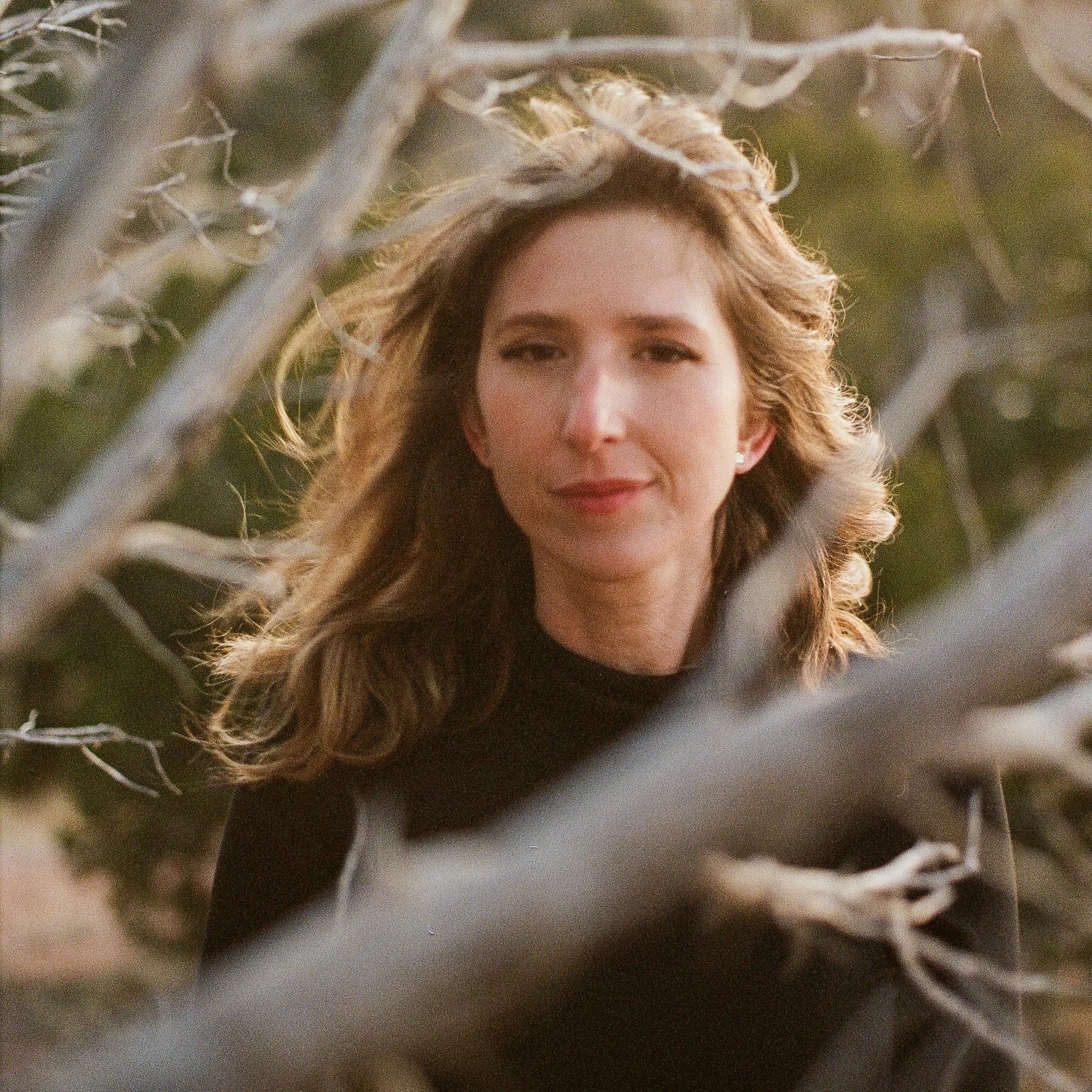 A woman with wavy light brown hair and fair skin. She is wearing a black top and is outdoors near some tree branches. The background is blurry with green and brown tones.