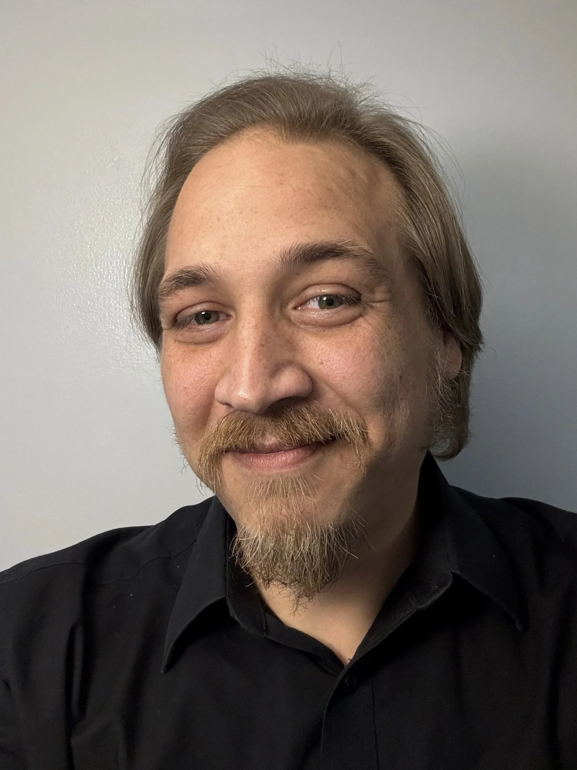 A man with light skin, light brown hair, a mustache, and a goatee smiling at the camera, wearing a black shirt, against a plain light gray background.