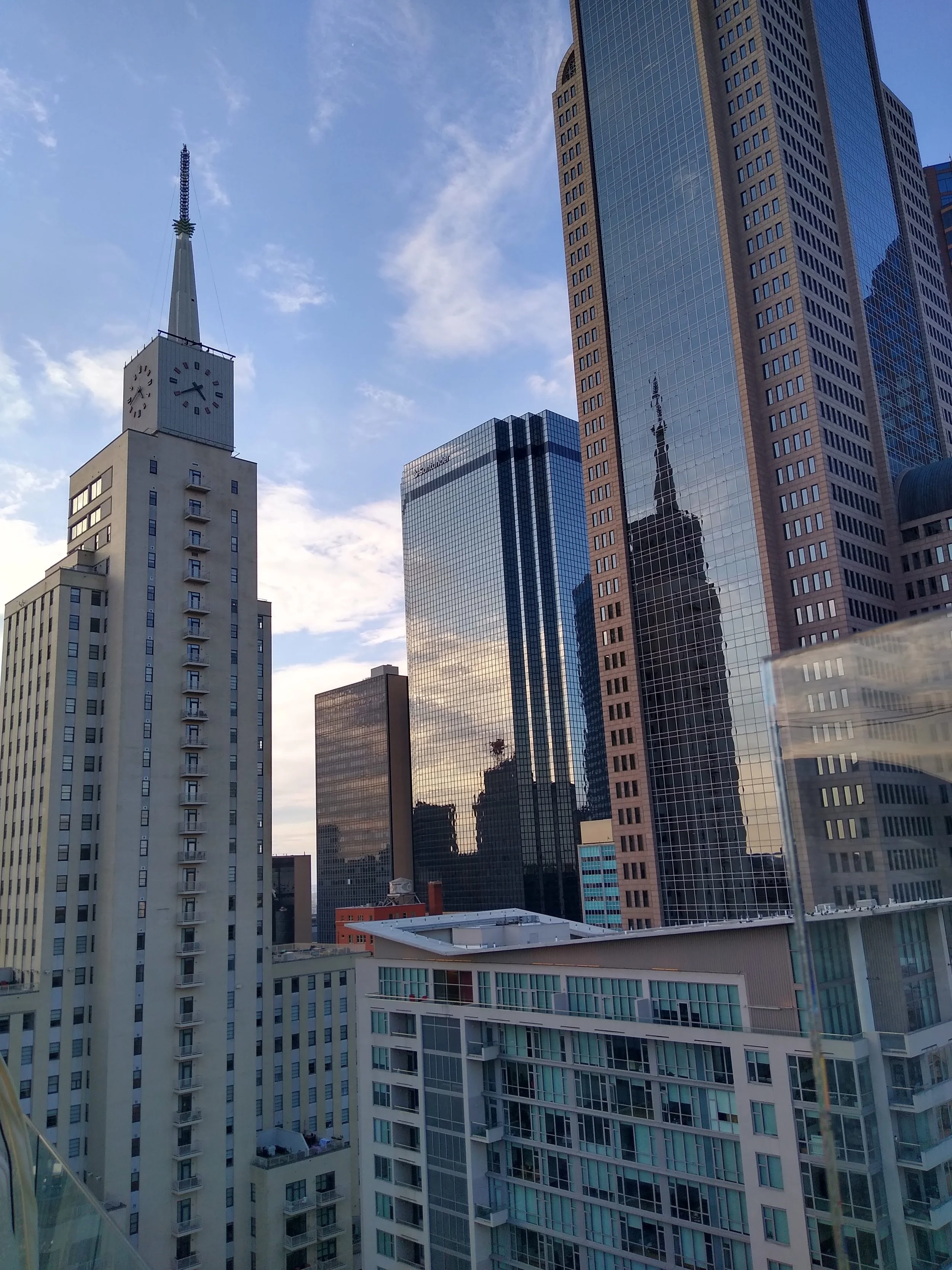 City skyline with tall skyscrapers, including the Century Plaza Tower with a clock and spire, and buildings with reflective glass facades reflecting other structures, under a partly cloudy sky.