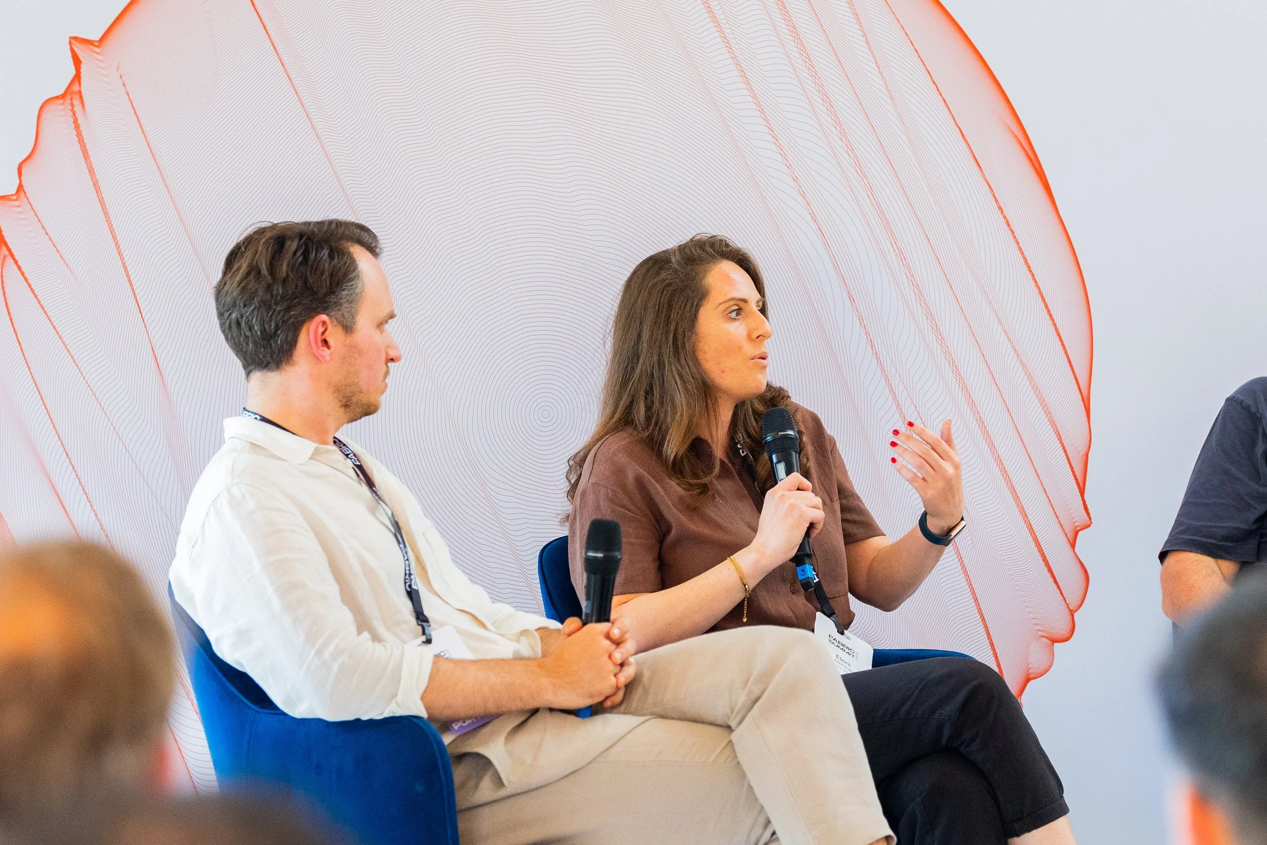 Two people, a man and a woman, speaking into handheld microphones during a panel discussion or conference. The woman is gesturing with her hand and has long brown hair, while the man has short brown hair and is wearing a white shirt. They are seated