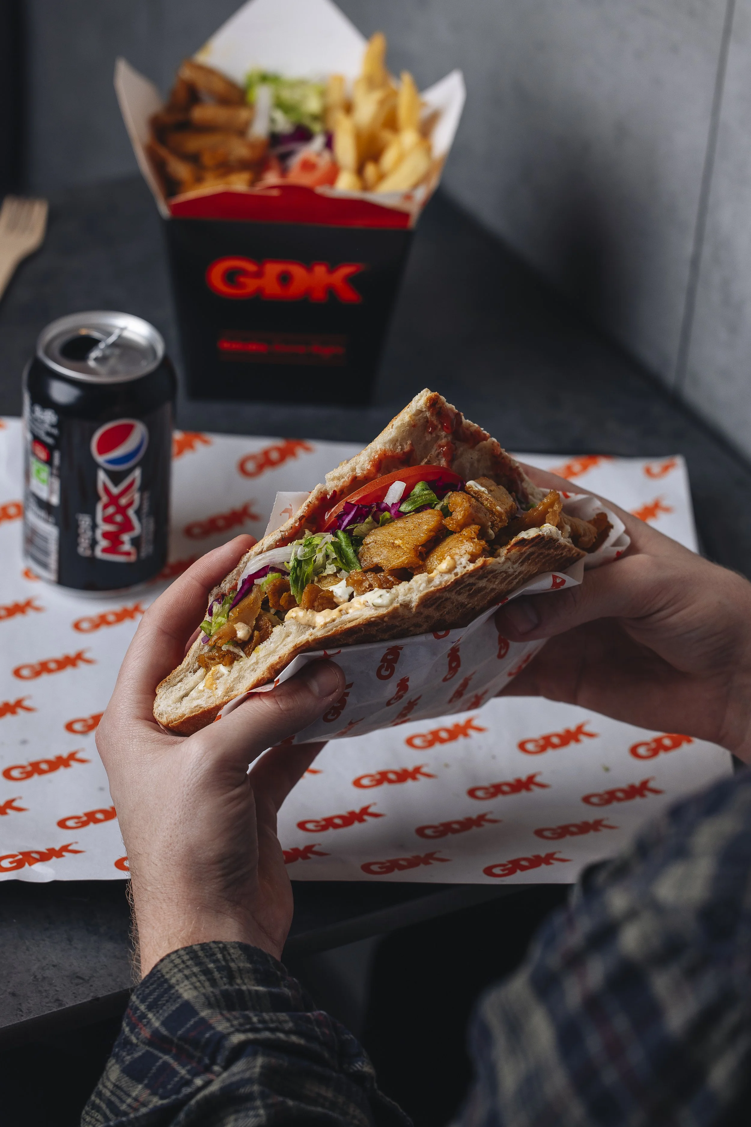 Person holding a sandwich with fries, vegetables, and meat, with a can of Pepsi and GDK food box on a table in the background.