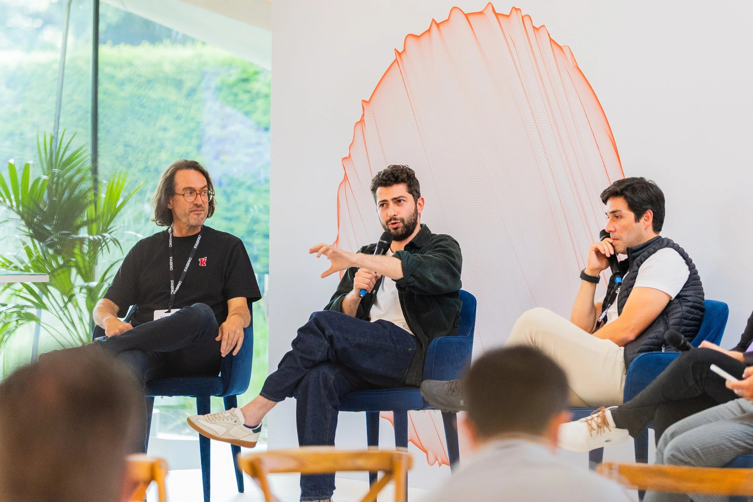 Three men participating in a panel discussion, one speaking with a microphone, in a modern indoor setting with artwork and plants.